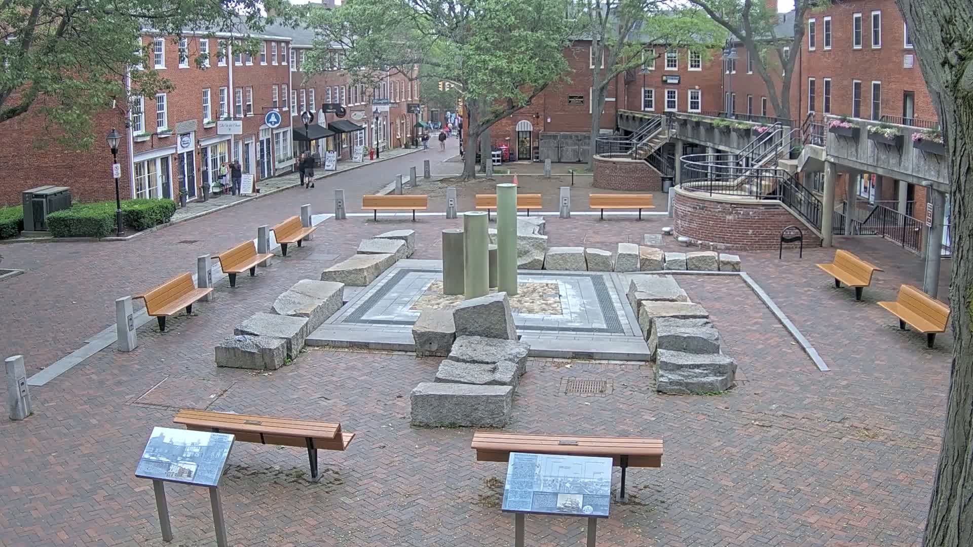 A brick plaza with several benches surrounds a stone fountain-like structure, under an overcast sky.