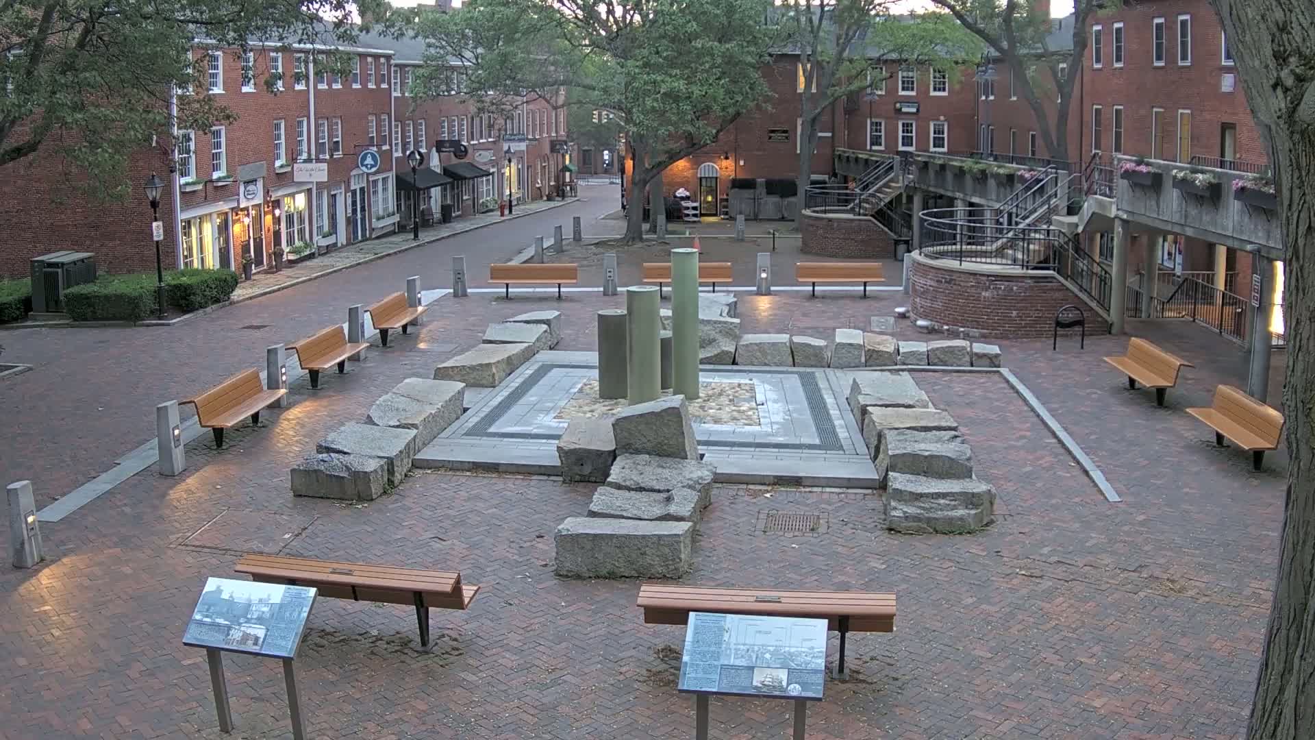 A brick plaza, containing a stone and metal fountain, is surrounded by benches and brick buildings under a partly cloudy sky.