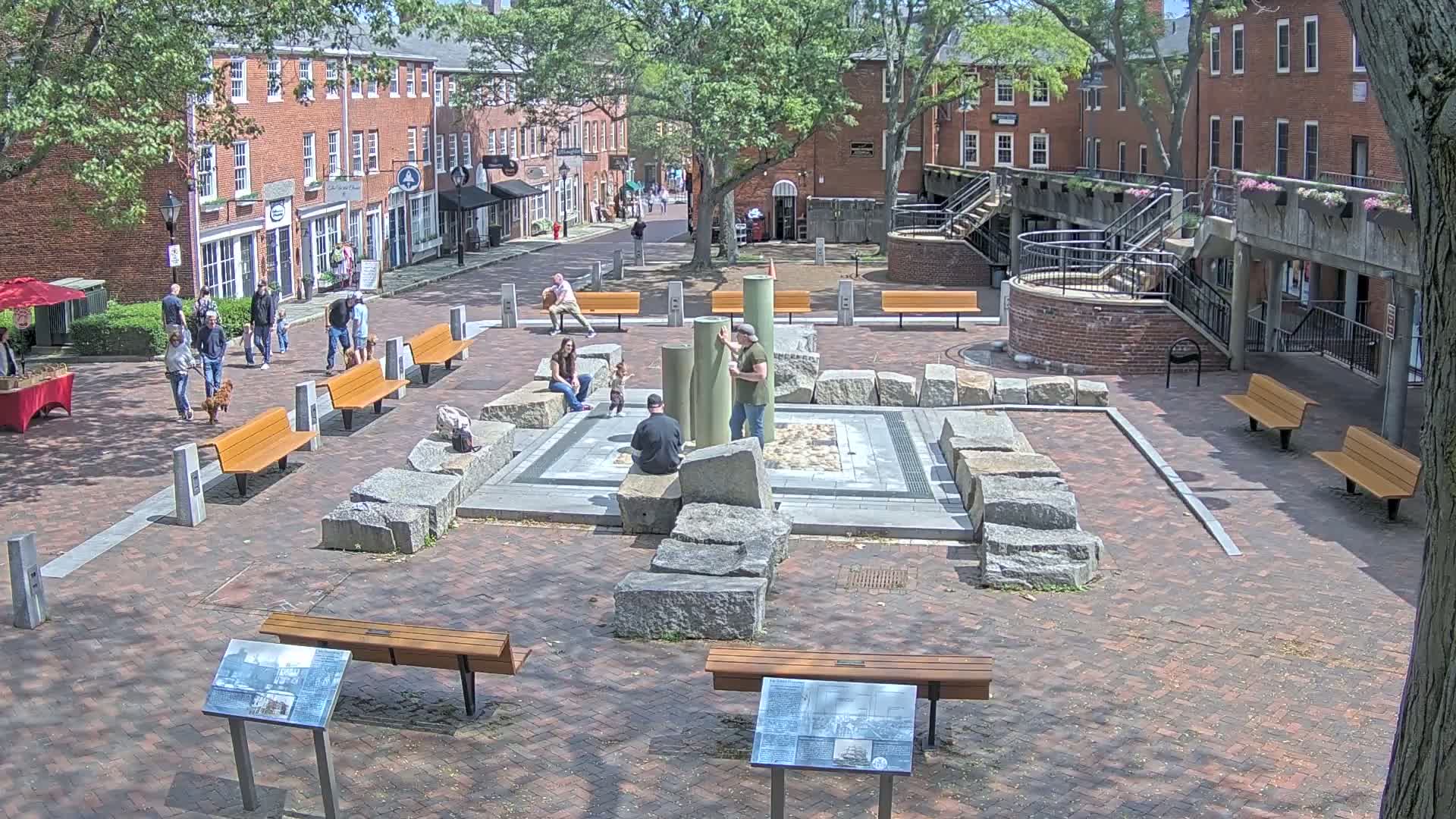A brick plaza with several benches, a stone fountain, and people walking around on a sunny day.