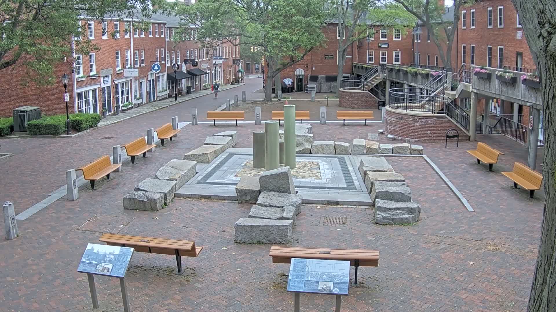 A brick plaza with several benches surrounds a central stone structure featuring upright cylinders, all under a partly cloudy sky.