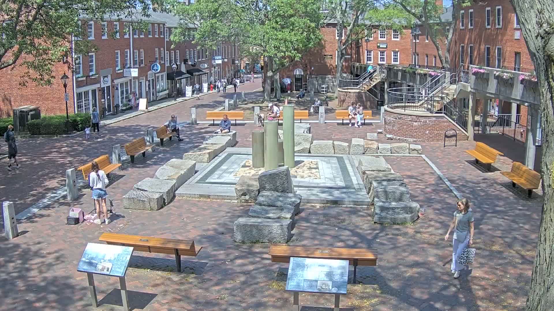 A sunny town square with brick buildings, benches, and a central stone fountain-like structure, has several people strolling and sitting.
