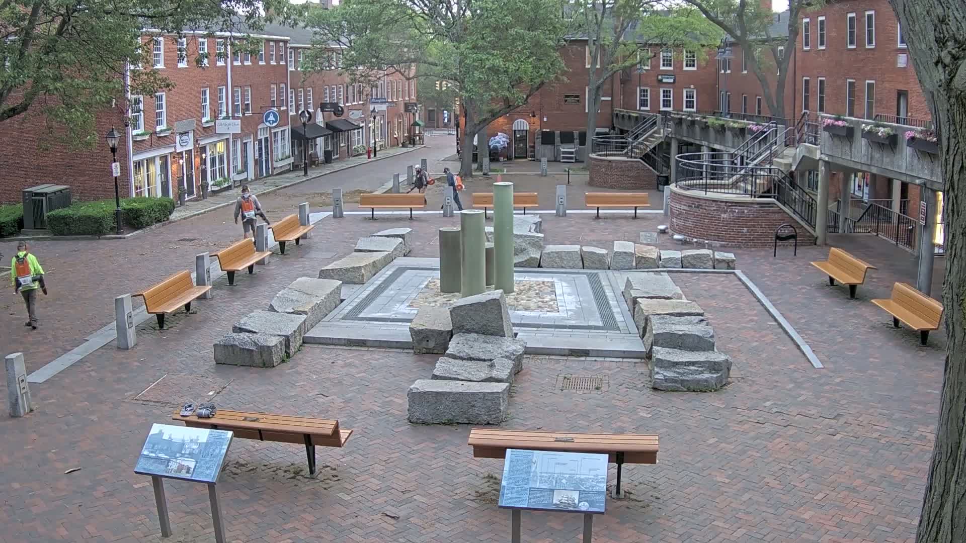 A brick plaza with several benches, a central stone structure, and surrounding brick buildings under an overcast sky.