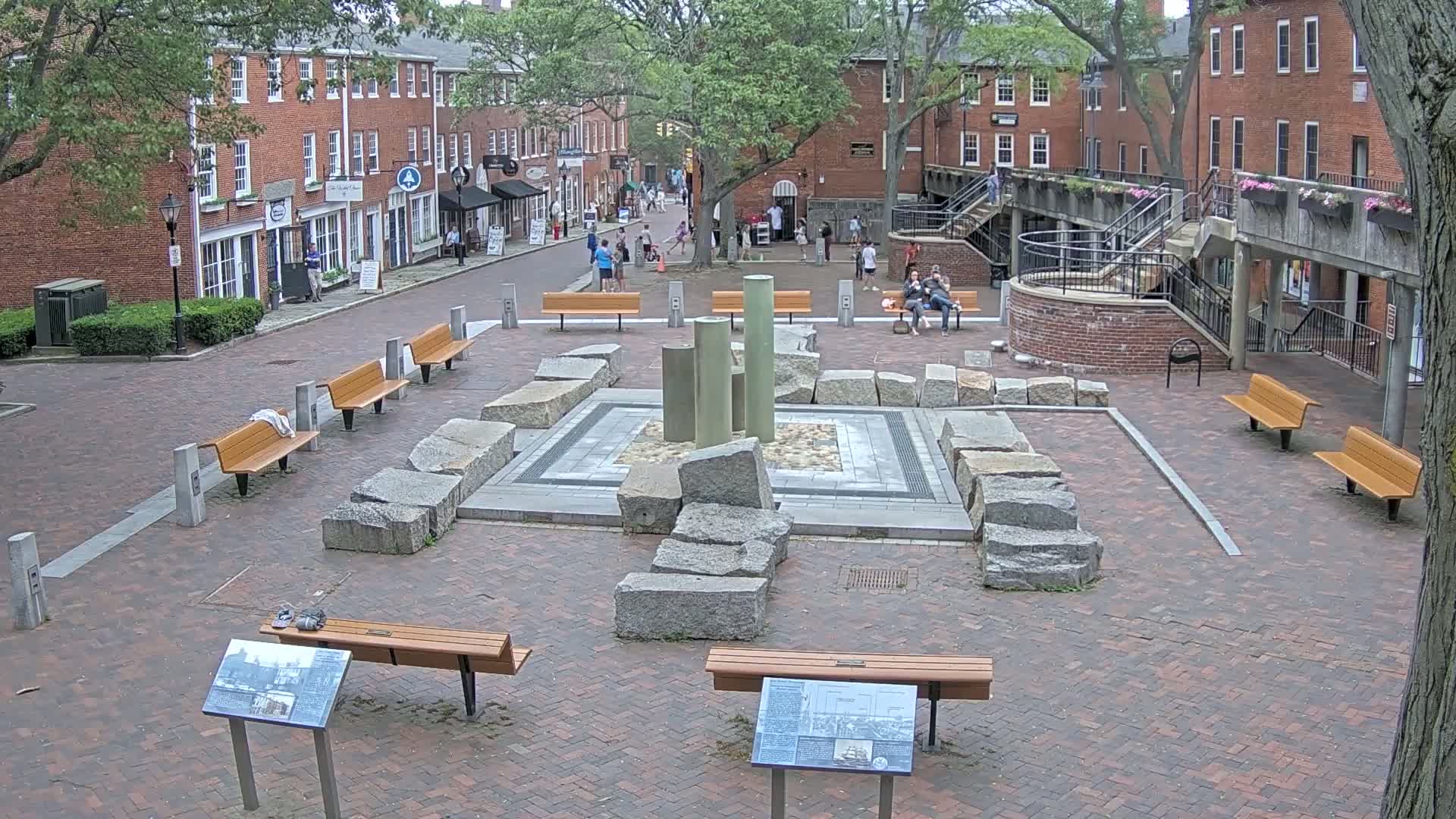 A brick plaza with benches, a central stone fountain, and several brick buildings in the background under an overcast sky.