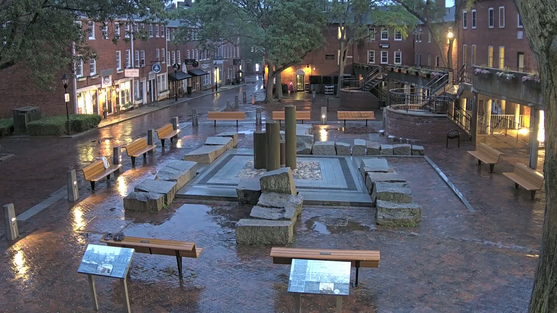 A rain-soaked brick plaza, featuring a central stone fountain and several benches, sits between brick buildings under a twilight sky.
