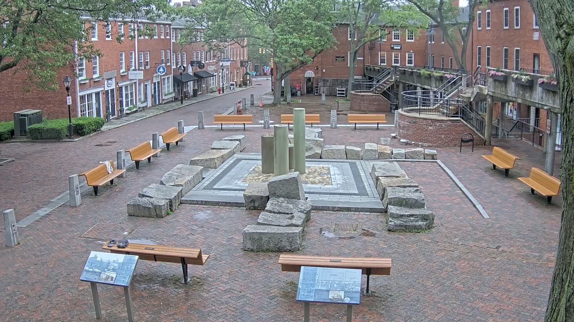A brick plaza with several benches surrounds a stone and metal fountain structure, situated between red brick buildings under an overcast sky.