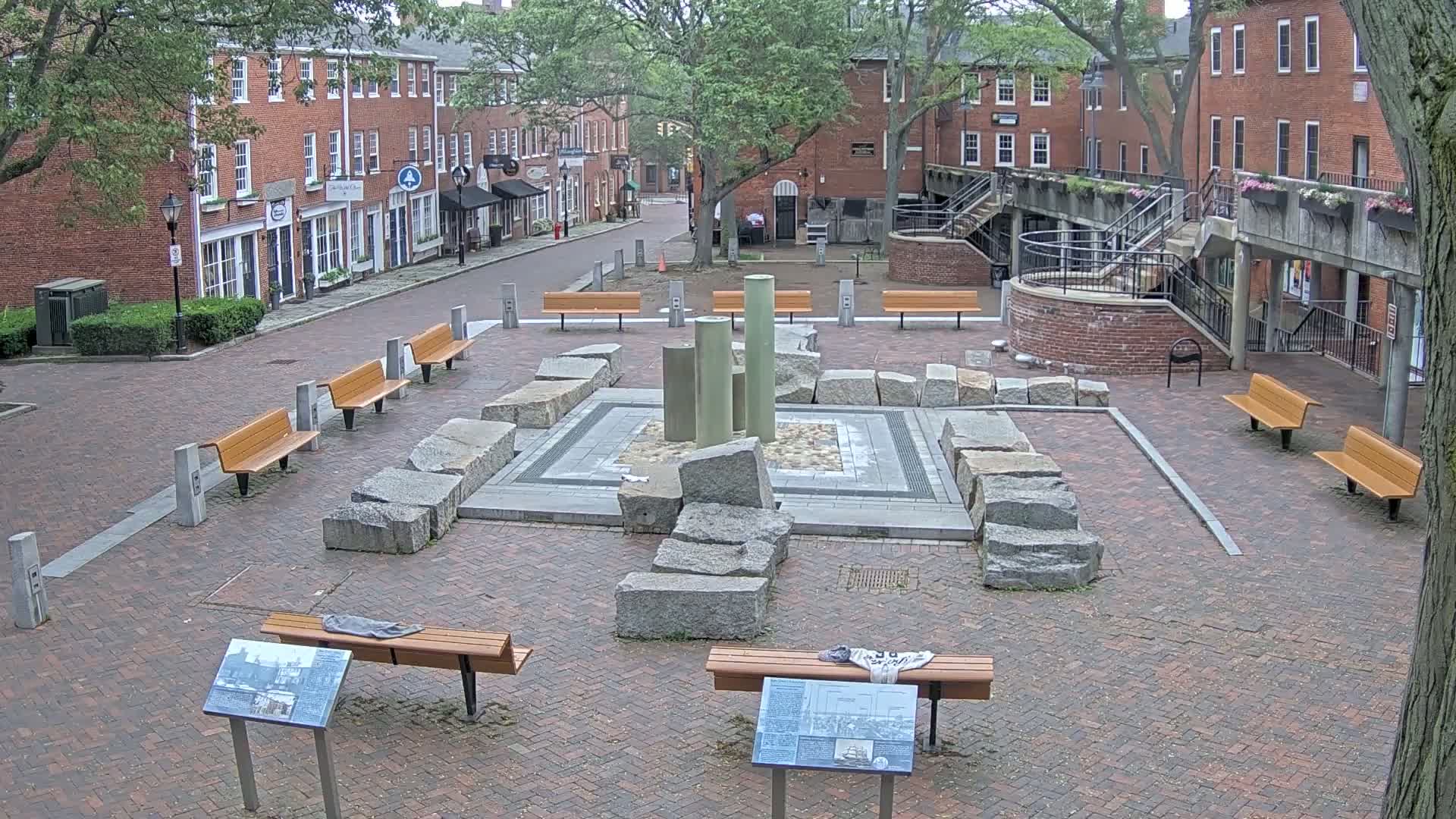 A brick plaza with benches surrounds a stone fountain;  red brick buildings line the street beyond.