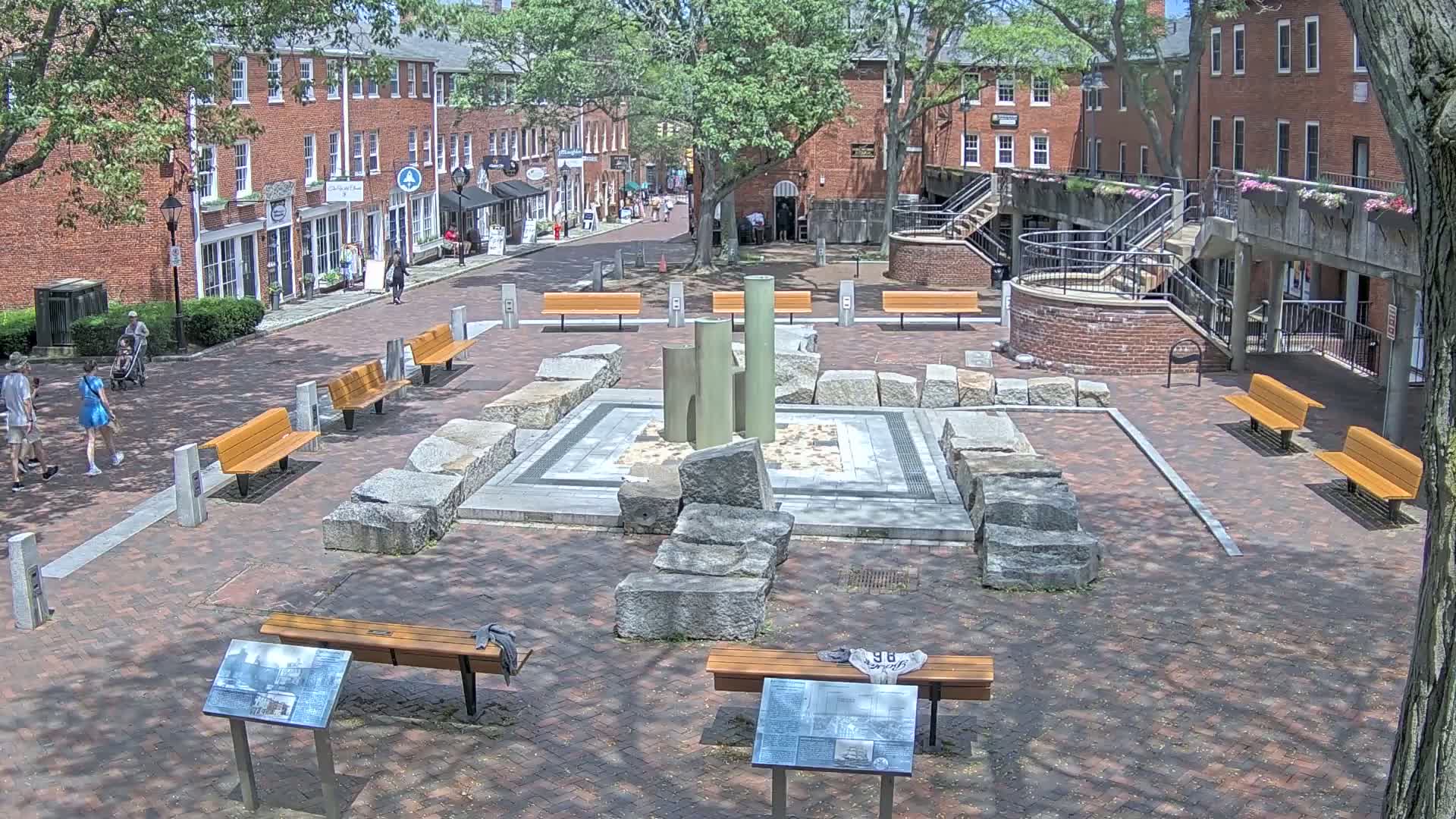 A brick plaza with several benches surrounds a central stone fountain structure, under a partly sunny sky.