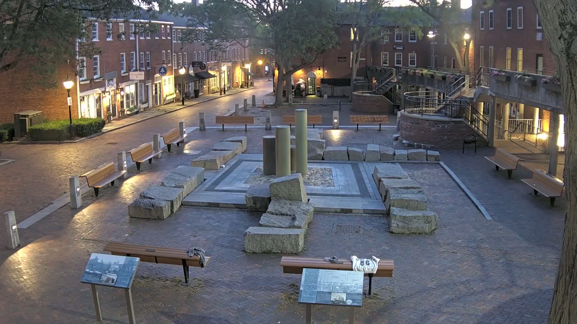 A brick town square at twilight features a stone monument with several pillars at its center, surrounded by benches and illuminated by streetlights.