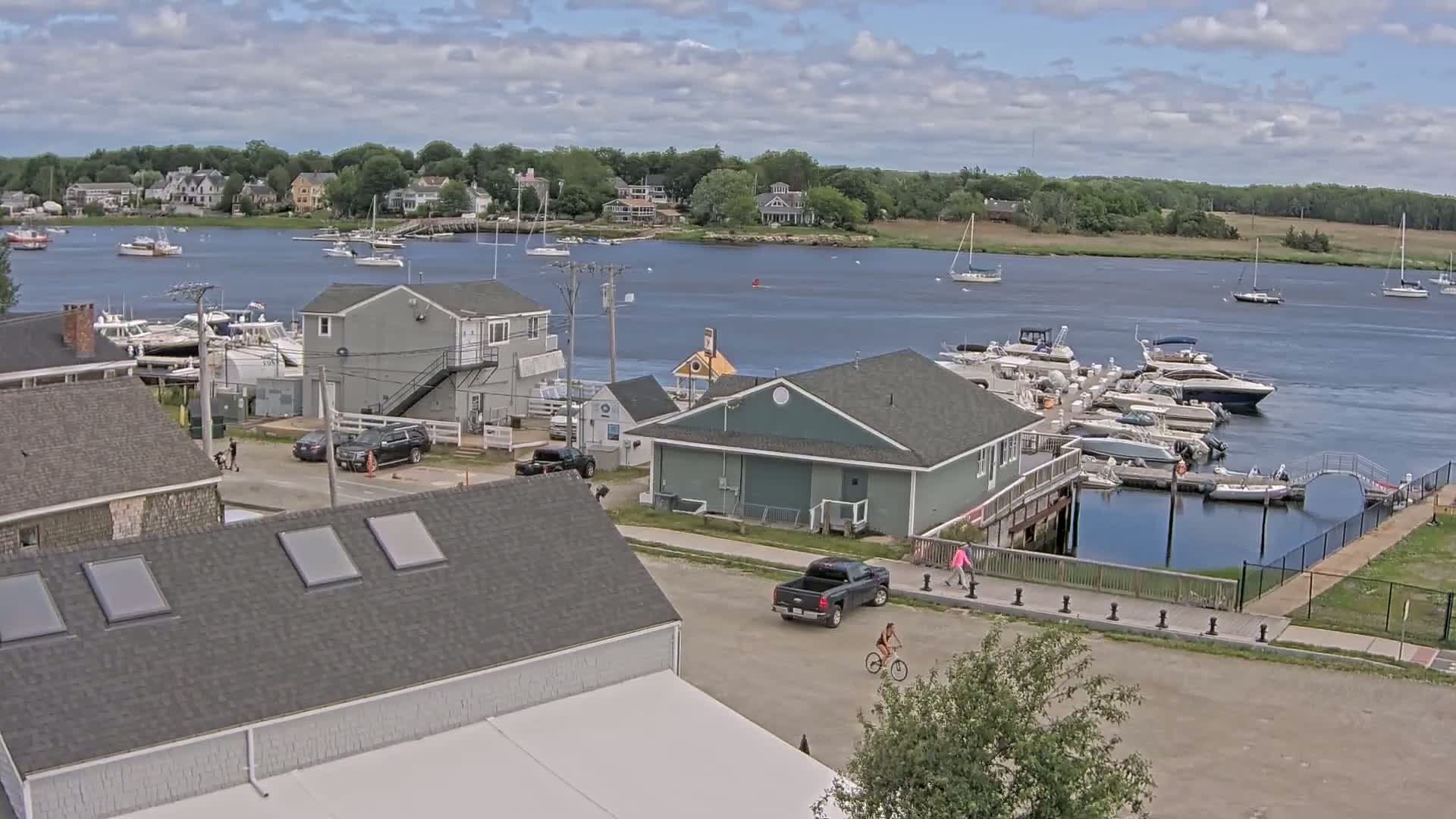 A waterfront scene on a partly cloudy day shows a marina filled with boats, several buildings, and a person riding a bicycle.