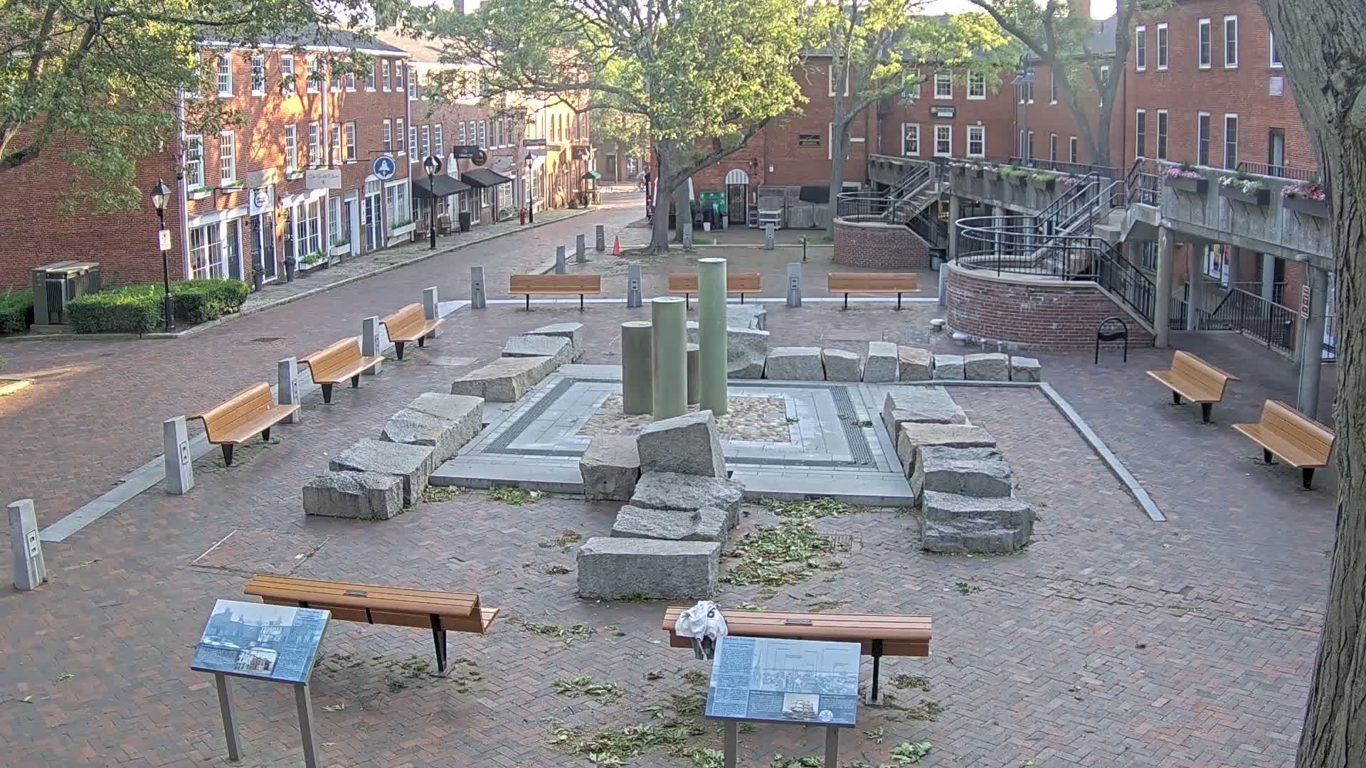 A brick plaza with several benches surrounds a stone structure and several pillars, all situated between brick buildings under a partly sunny sky.