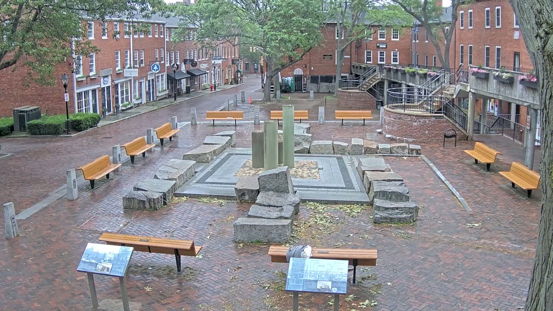 A brick plaza, partly wet from rain, features a central stone structure with several benches and informational signs around it, bordered by red brick buildings with green trees.