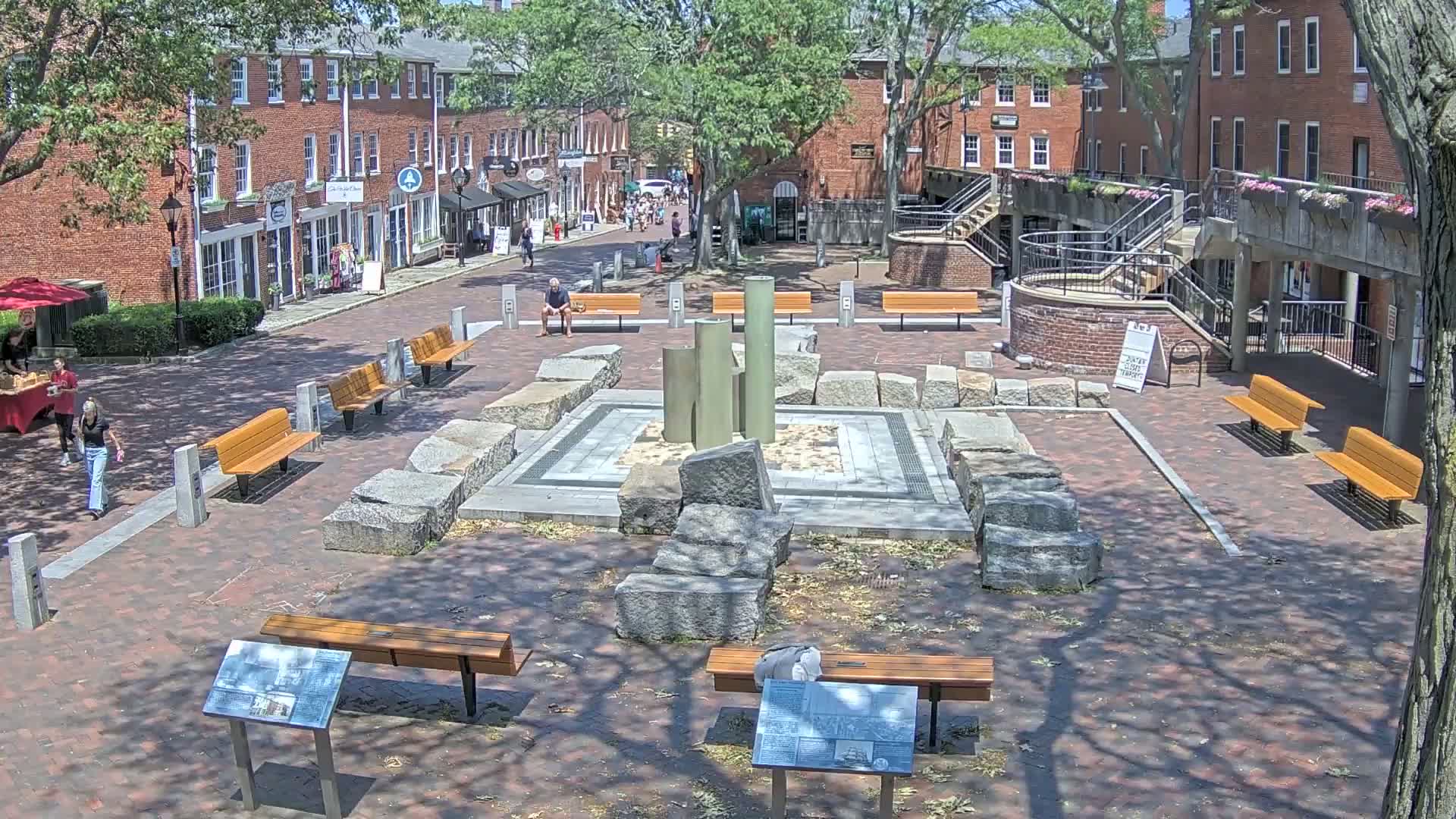A brick plaza with benches and a stone fountain is surrounded by brick buildings on a sunny day.