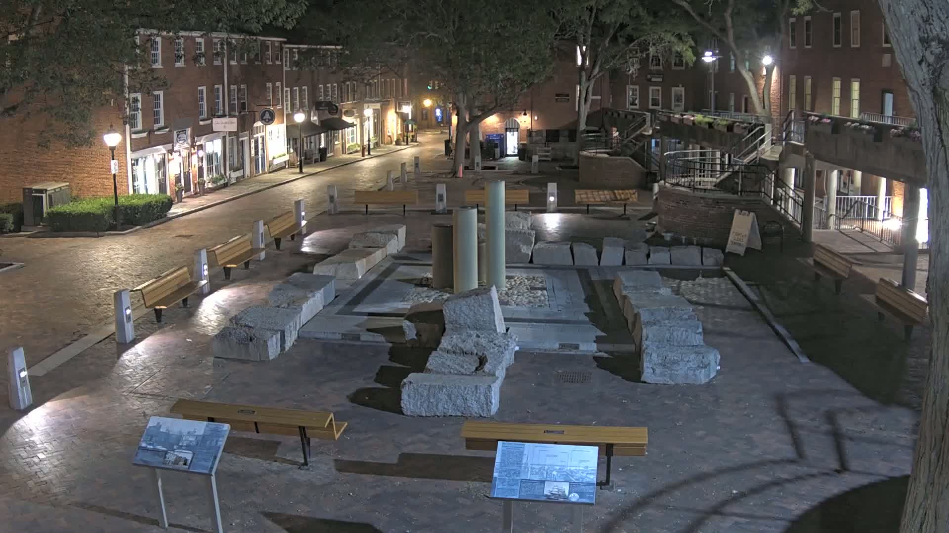At night, a brick-paved street lined with brick buildings leads to a square containing large stone remnants and several benches under the shade of trees.