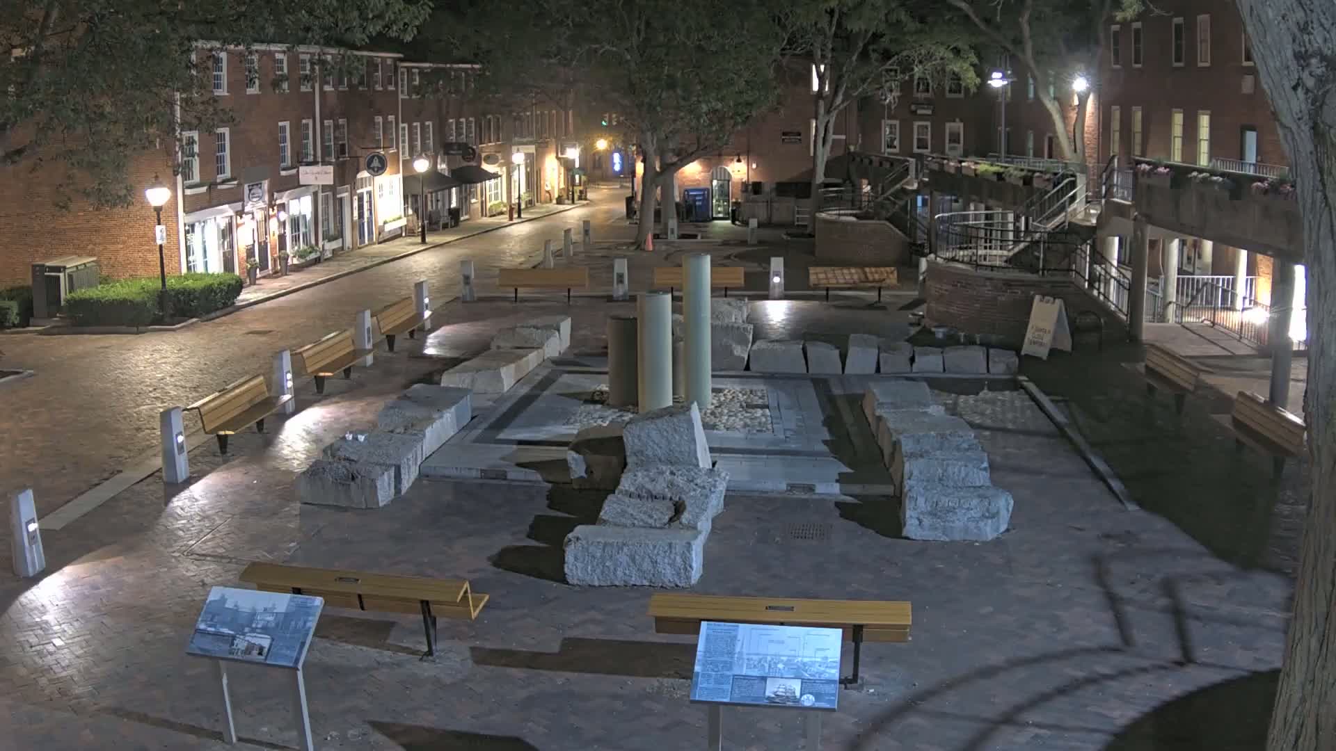 At night, a brick-paved town square contains a low stone structure with several large stone blocks and three upright columns, surrounded by benches and lit by streetlights.