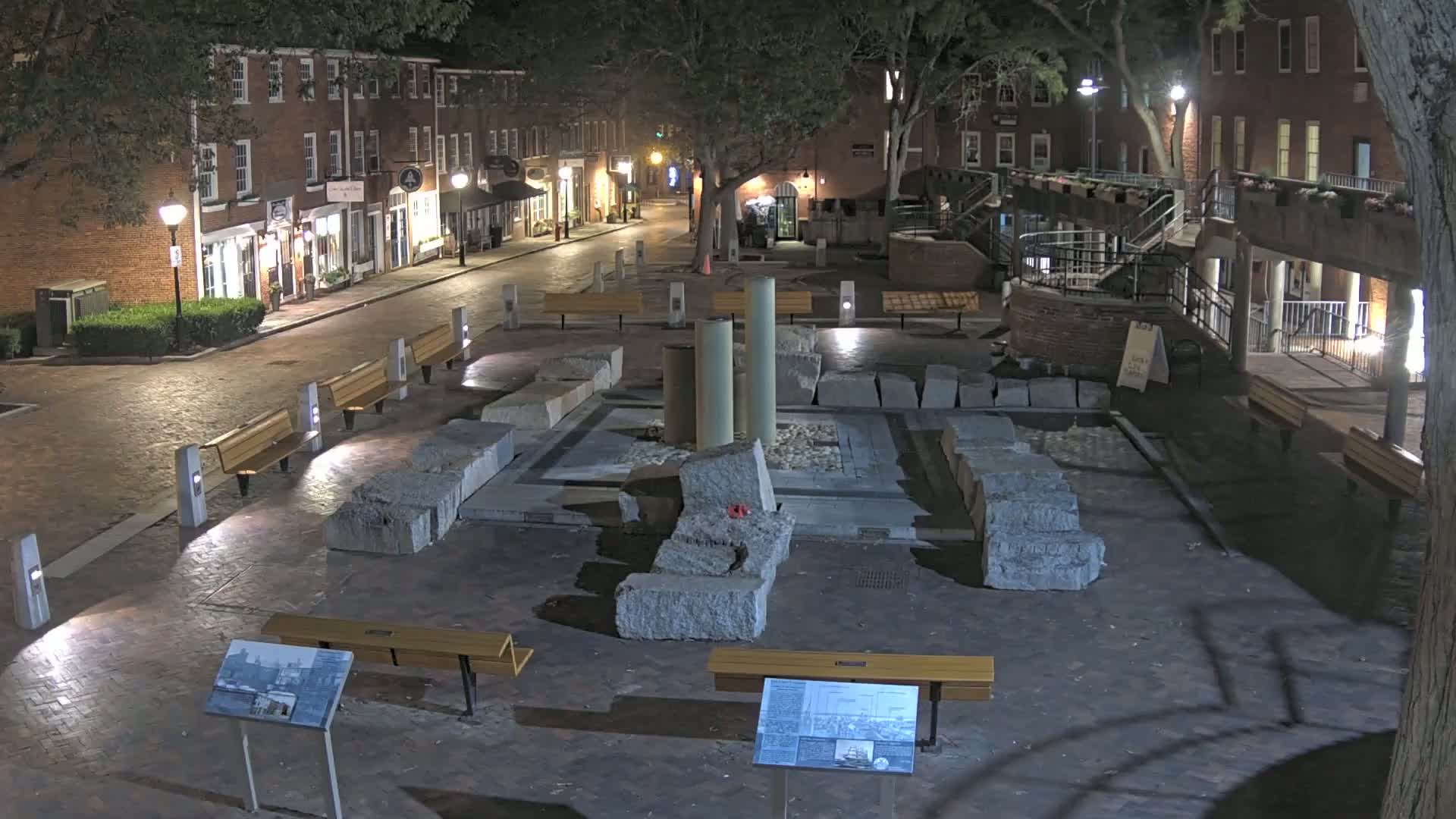 At night, a brick street lined with buildings leads to a plaza containing large stone blocks, several benches, and three tall pillars.