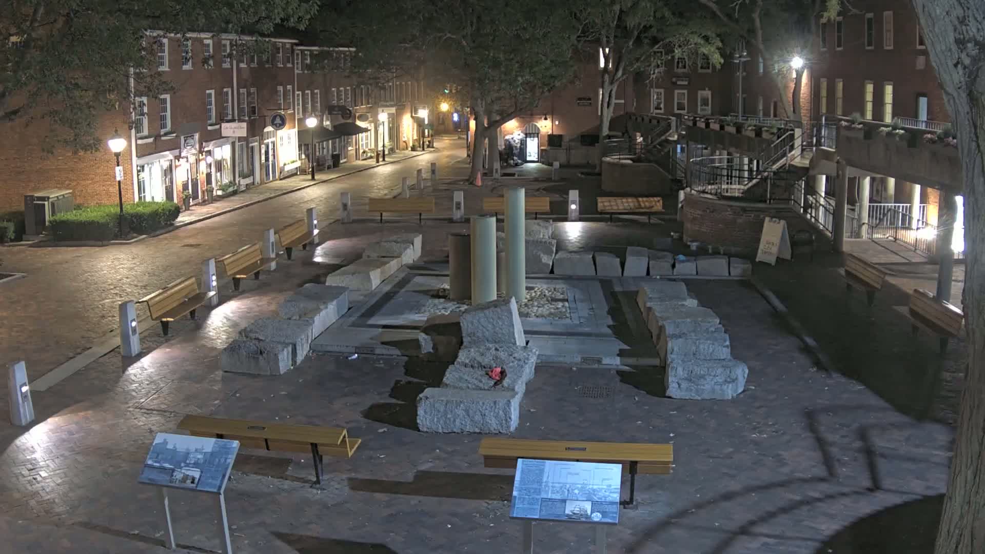 At night, a stone plaza with benches and informational signs sits between brick buildings under the cover of trees.