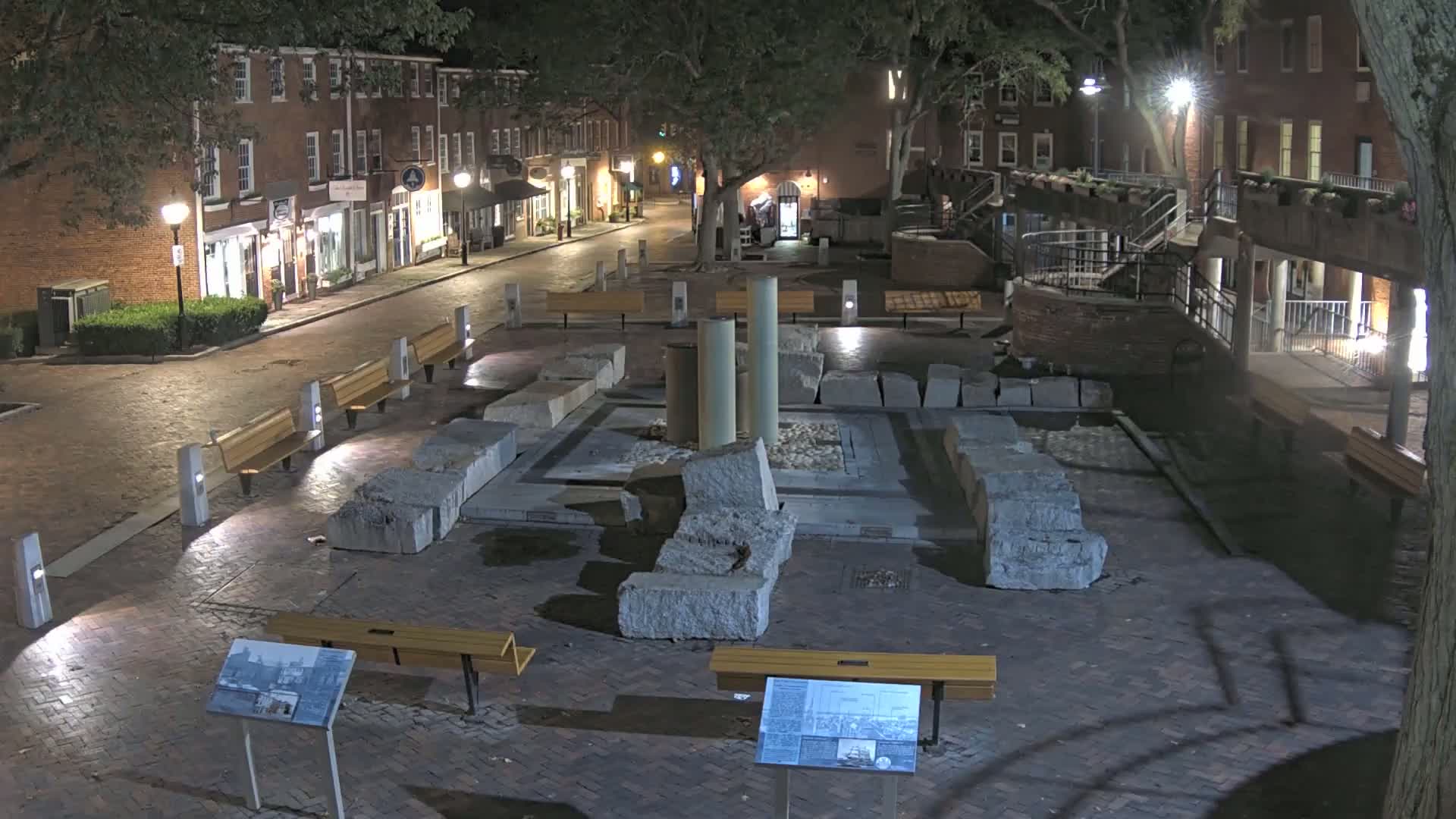 A nighttime view of a brick plaza with large stone ruins at its center, surrounded by brick buildings and park benches under the cover of trees.
