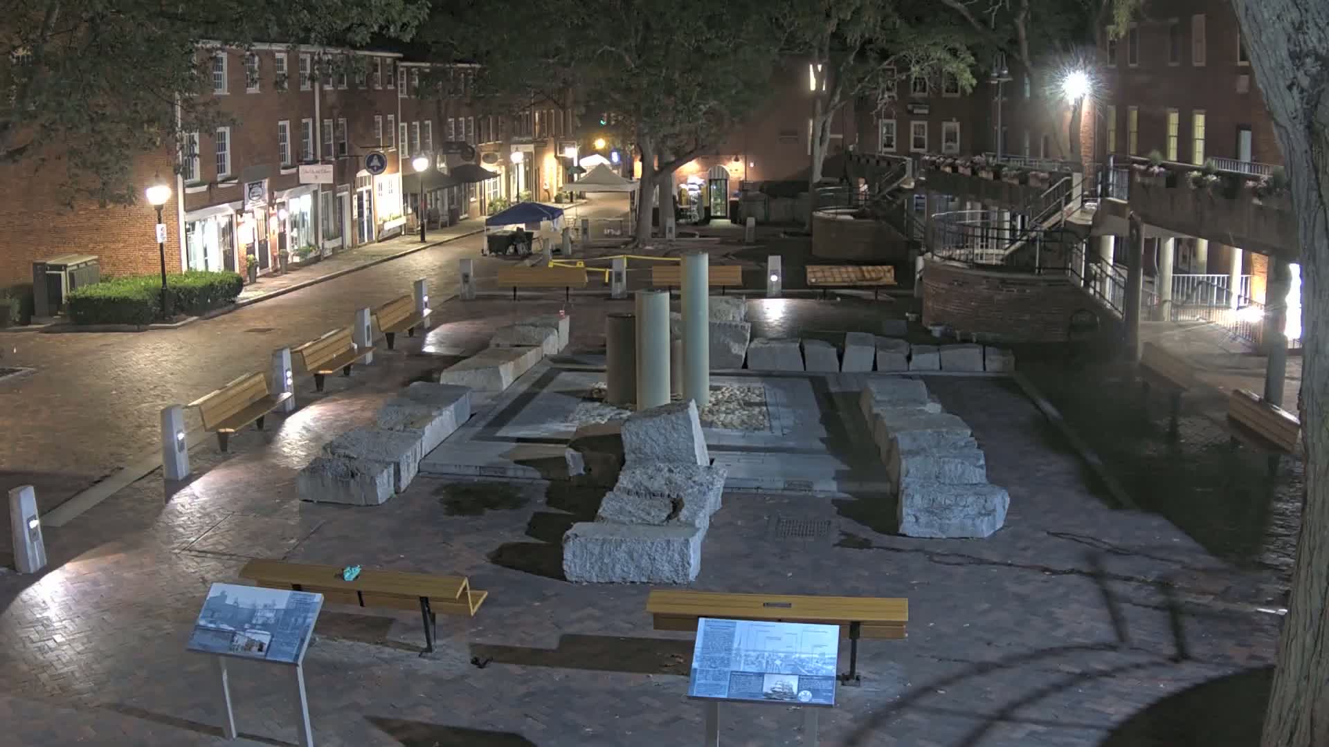 A nighttime view of a brick-paved town square featuring a stone structure, benches, and streetlights, with brick buildings lining a street in the background.