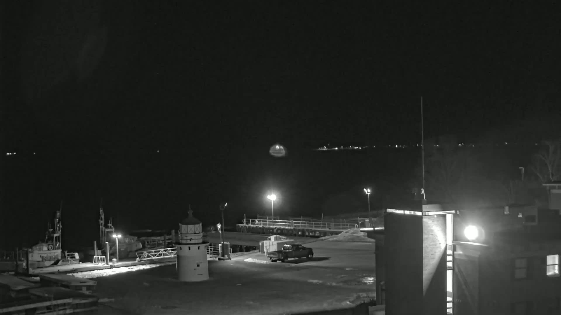 A nighttime, black-and-white view of a harbor with a lighthouse, boats, a truck, and a building under a dark sky.