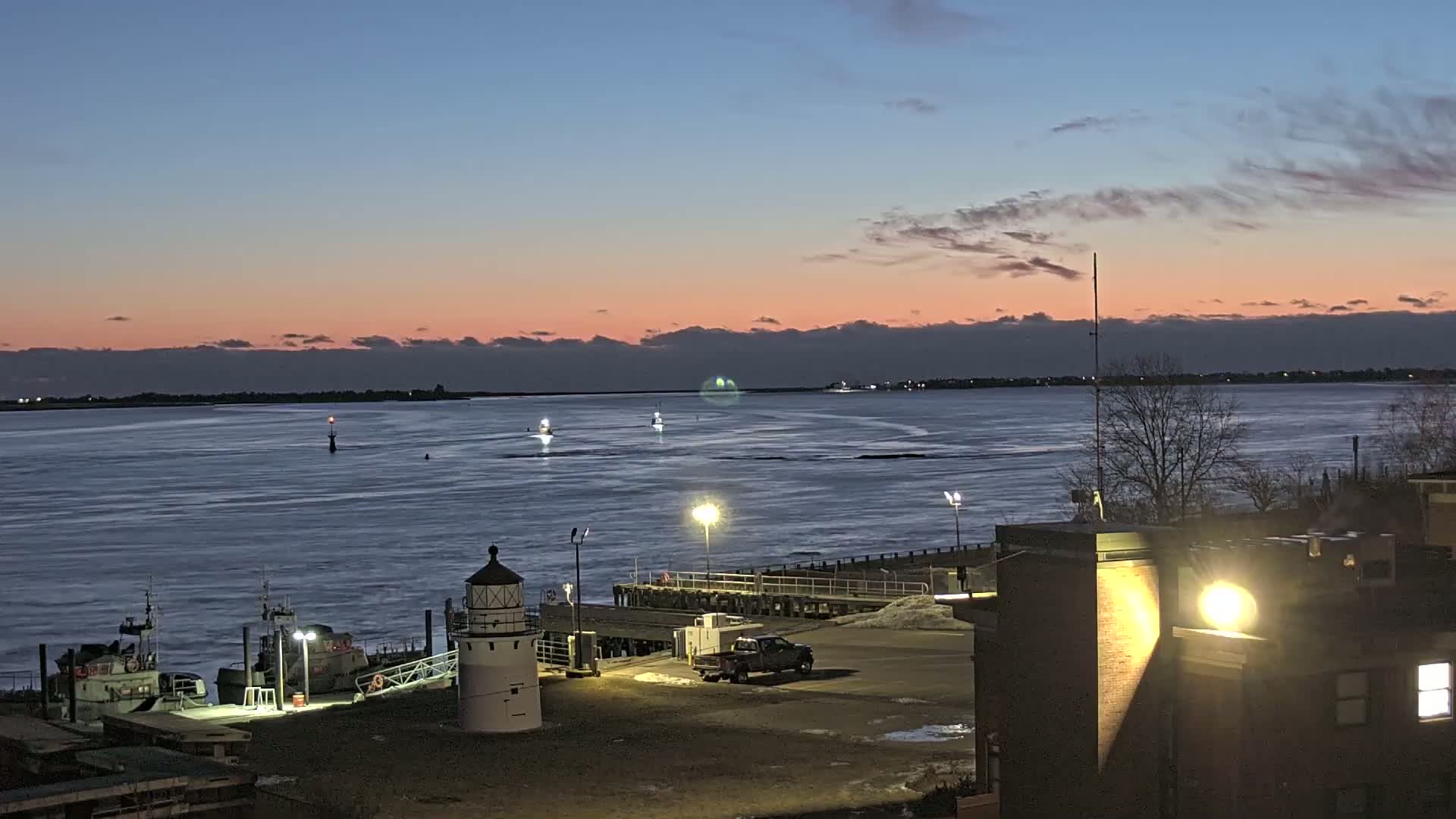 A twilight view of a harbor with a lighthouse, boats, a pier, and a building, under a partly cloudy, pinkish sky.