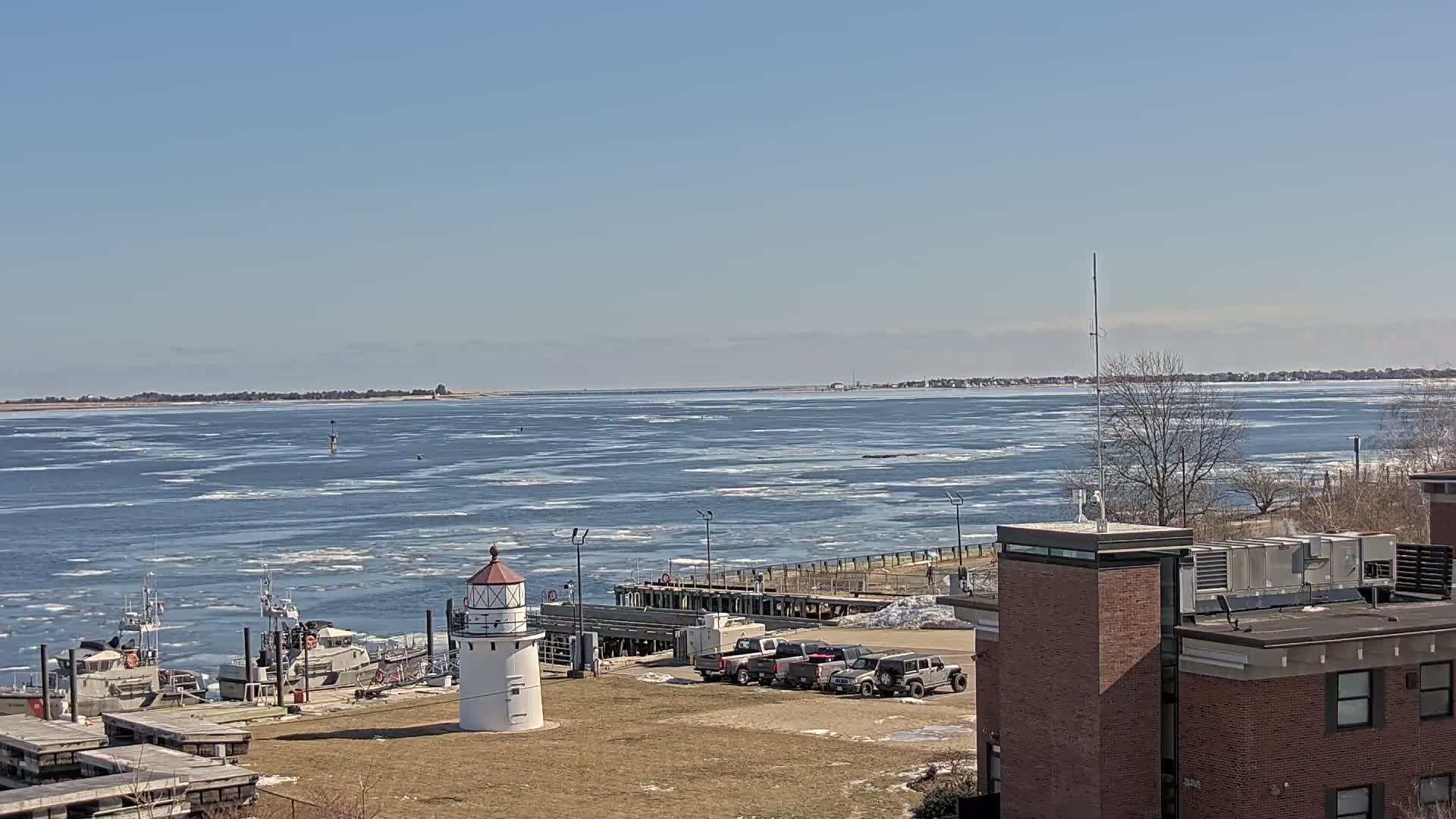 A partially ice-covered body of water is seen from a distance, with several boats docked near a small lighthouse and a building on a clear, sunny day.
