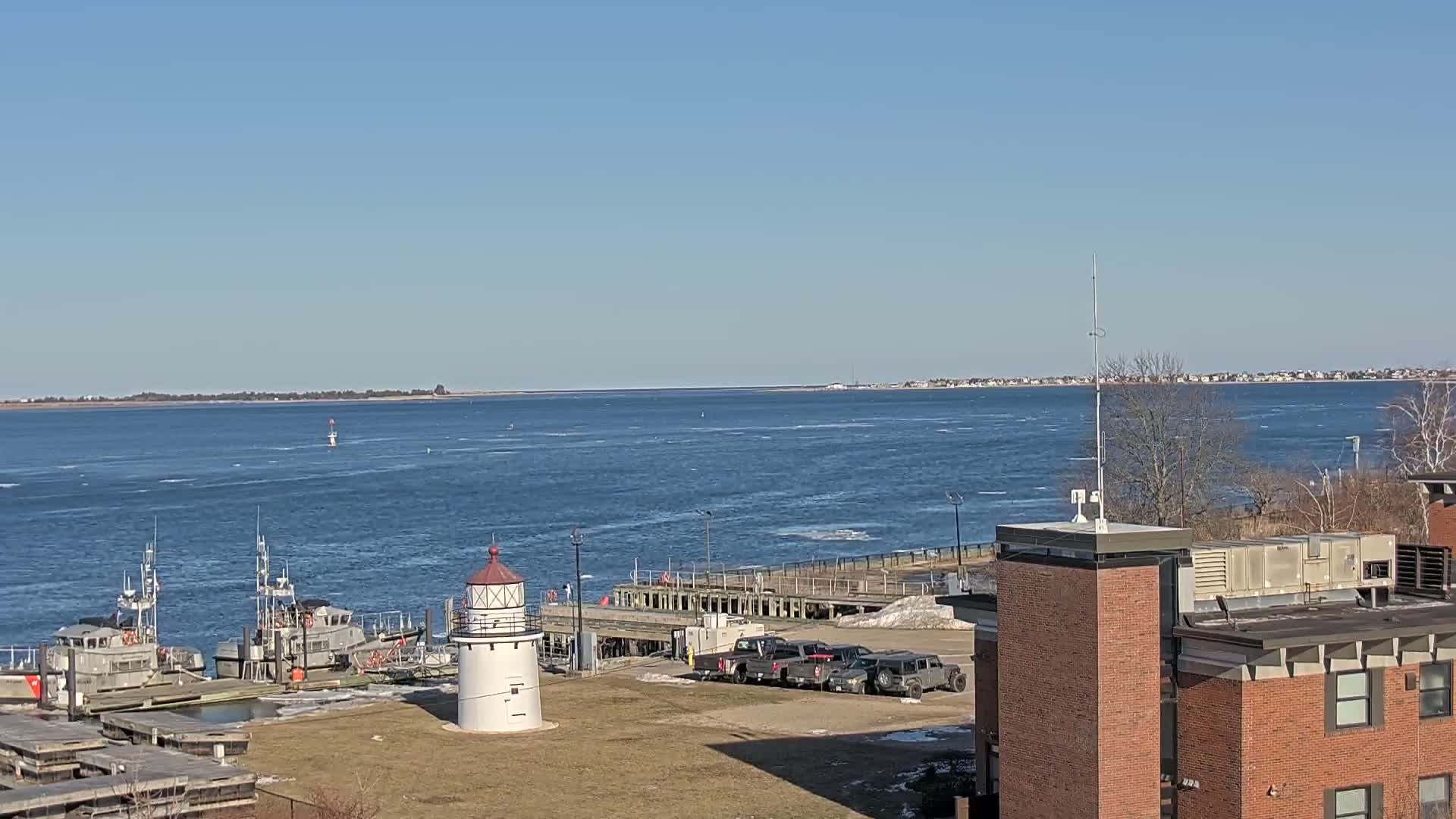 A small white lighthouse stands near a dock with several boats and a few vehicles, overlooking a body of water with a distant shoreline under a clear, sunny sky.