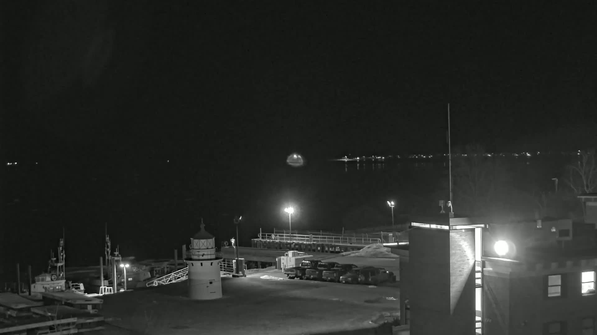 A nighttime view of a harbor with a lighthouse, boats, and parked cars under a dark, clear sky.