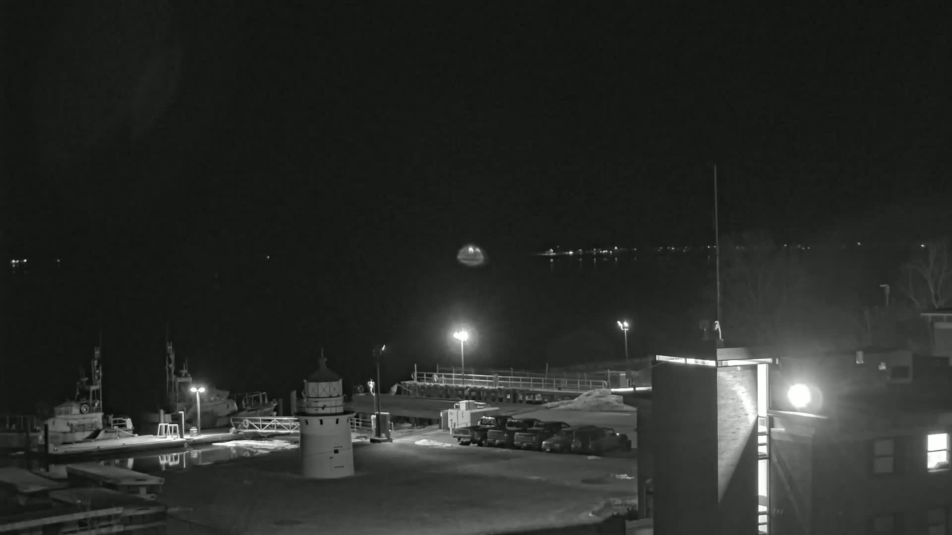 A nighttime view of a harbor with boats docked, a lighthouse, and a few parked cars, under a dark sky.