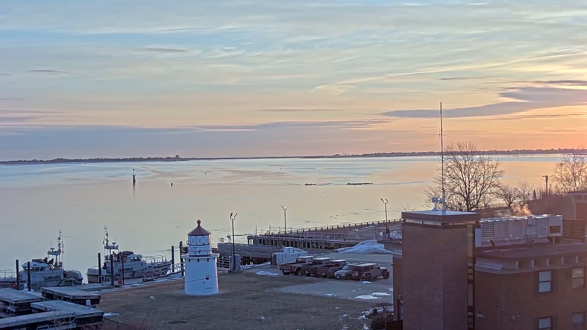A calm, pale-pink sunrise illuminates a harbor scene with a small lighthouse, docked boats, and a few parked vehicles near a building.