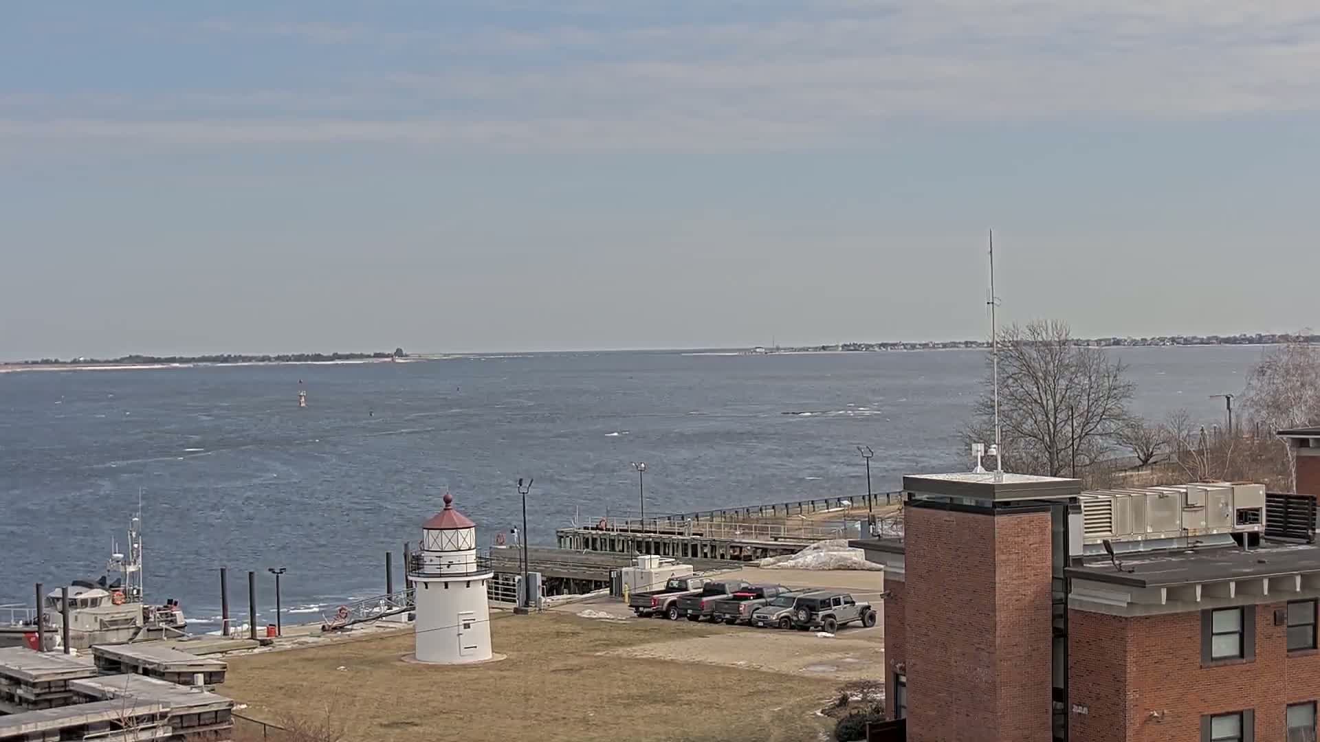 A small lighthouse stands near a dock and a body of water under a mostly clear sky, with several vehicles parked nearby and a building in the foreground.