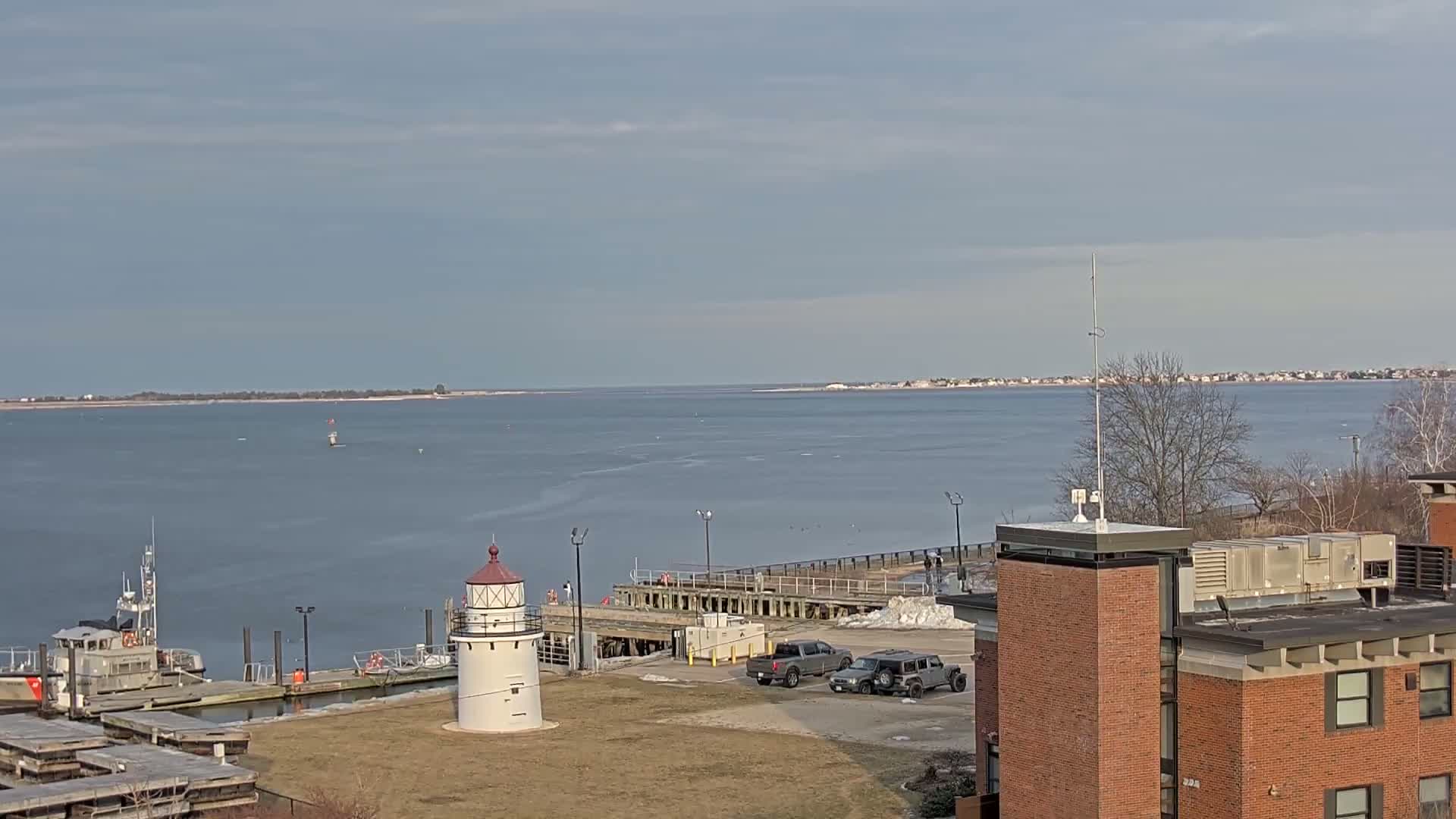 A white lighthouse stands near a dock and a Coast Guard vessel, overlooking a calm body of water under a mostly cloudy sky.