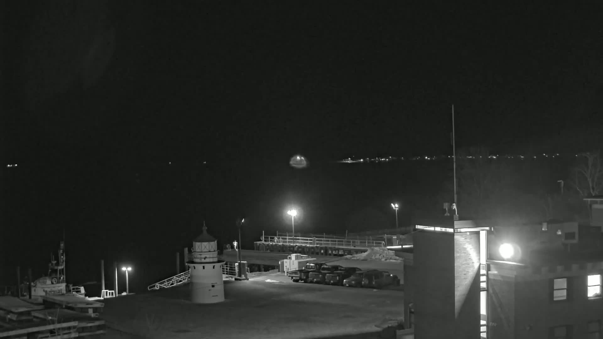 A nighttime, black-and-white view of a harbor with a lighthouse, several parked vehicles, and a building, under a dark, clear sky.
