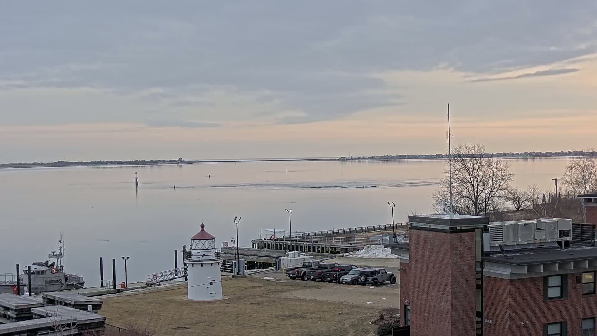 A white lighthouse stands on a grassy area near a calm body of water under a pale, cloudy sky, with a small boat docked nearby and several vehicles parked in a lot behind it.