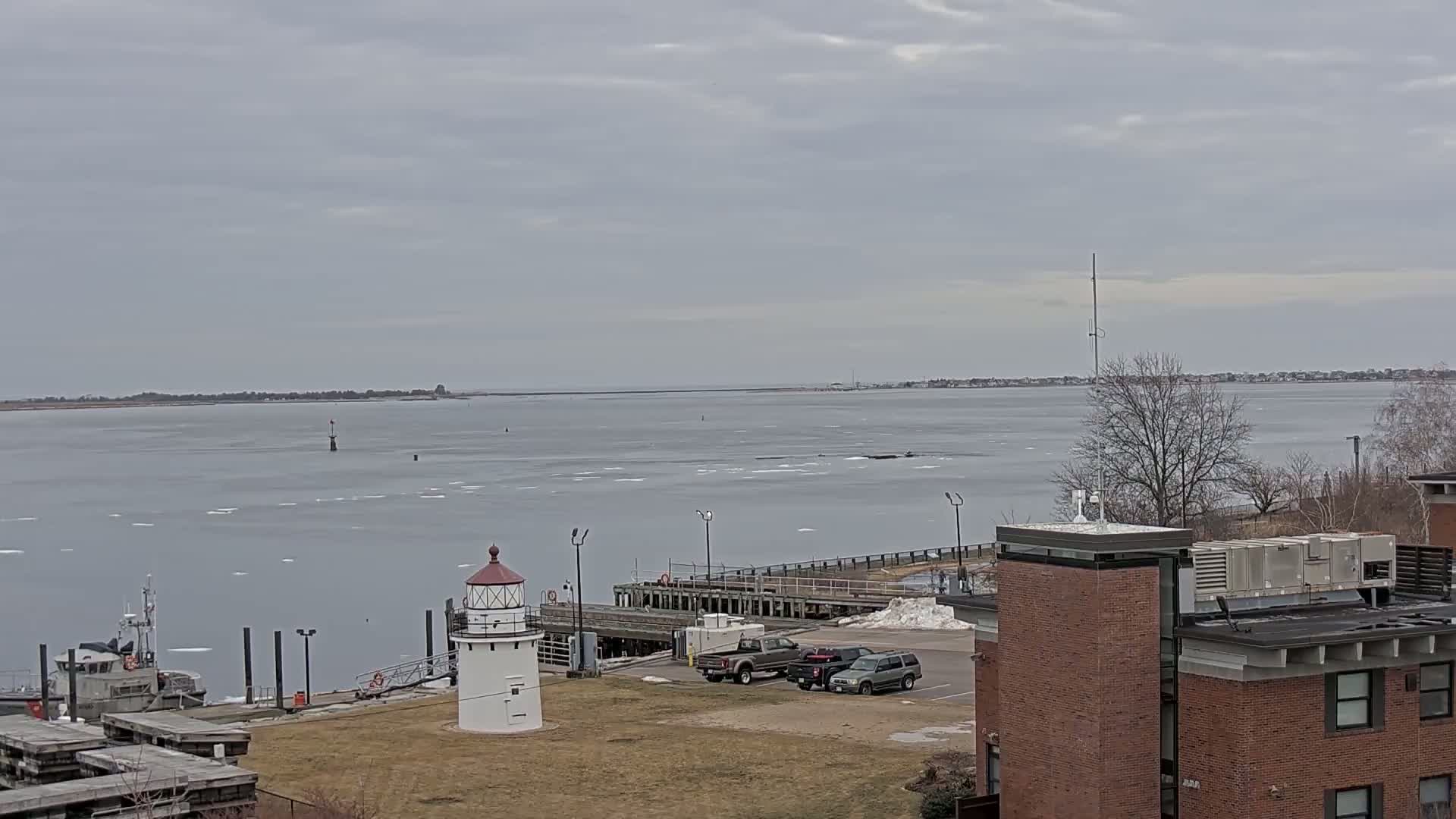 A small lighthouse stands on a grassy area overlooking a partially ice-covered bay on an overcast day, with a small boat and several vehicles visible nearby.