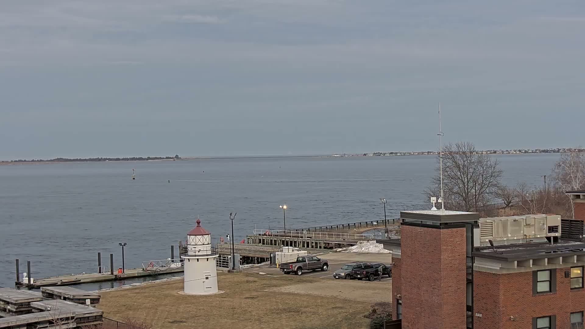 A small lighthouse stands on a grassy area next to a dock and body of water with a distant shoreline under an overcast sky.