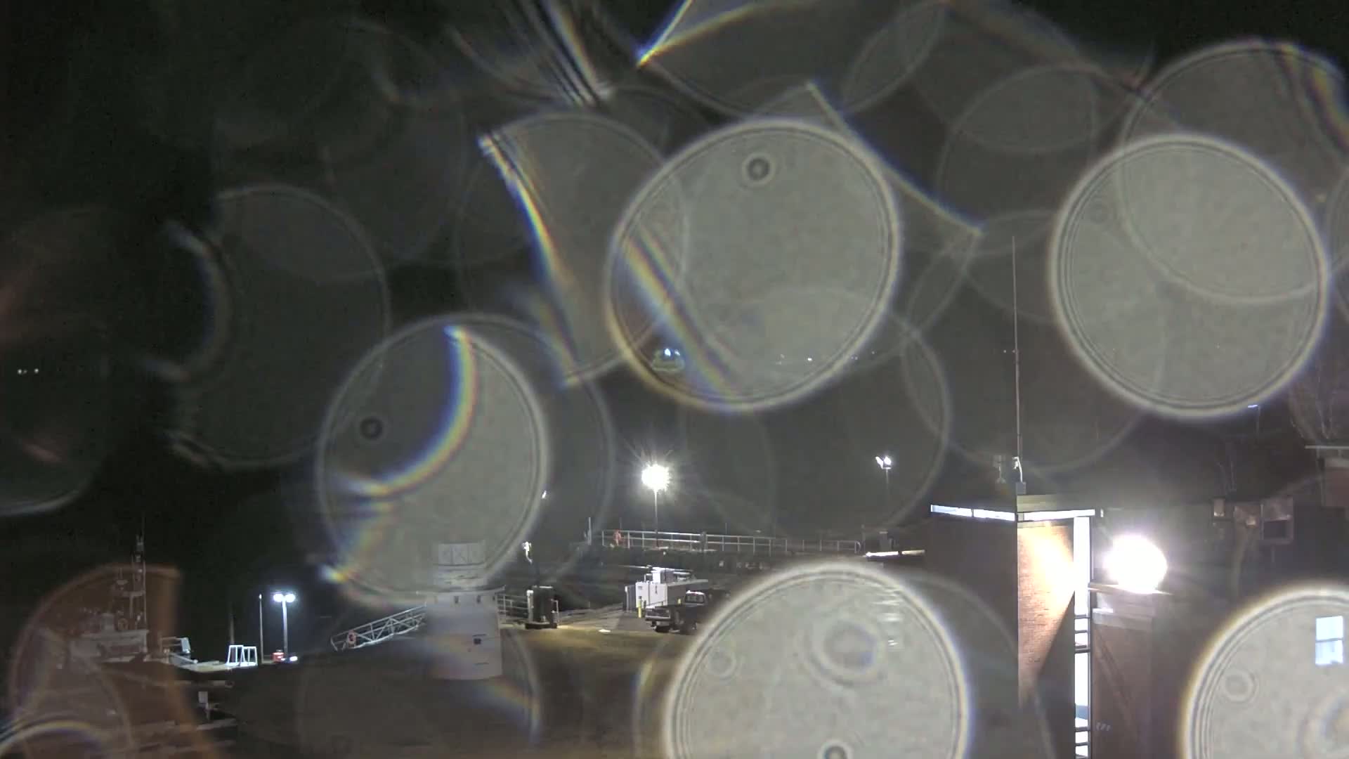 A nighttime view of an industrial area with several buildings and vehicles, seen through a rain-covered window causing blurry, circular distortions with rainbow-like effects.
