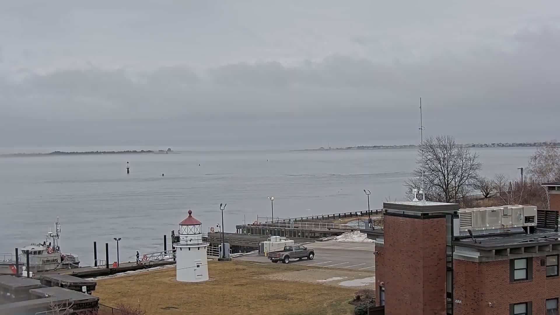 A small lighthouse stands near a dock and a small boat on a calm, overcast day overlooking a body of water and distant land.