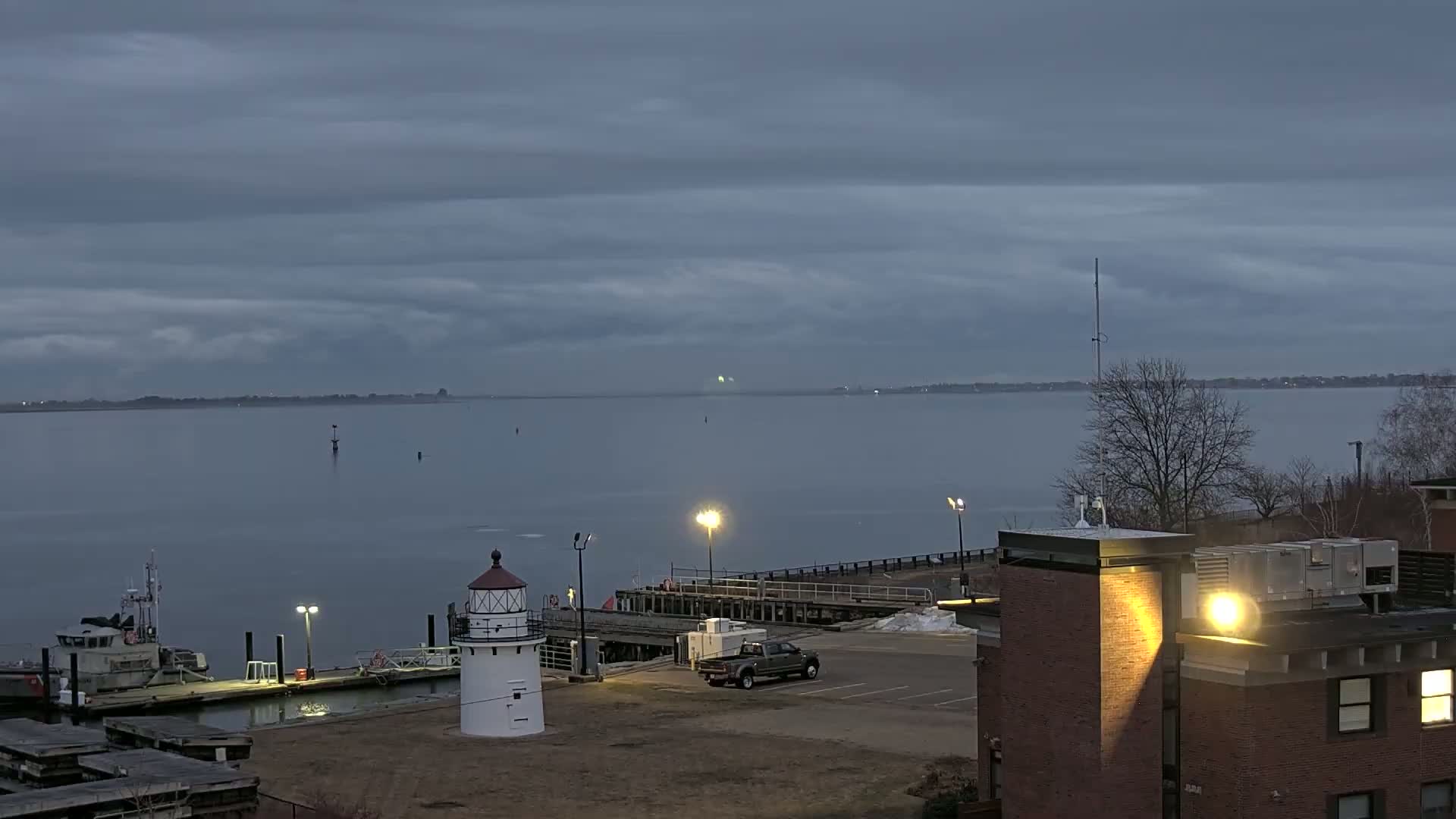 A small lighthouse stands near a docked boat and a truck in a parking lot overlooking a calm, gray body of water under a cloudy sky.