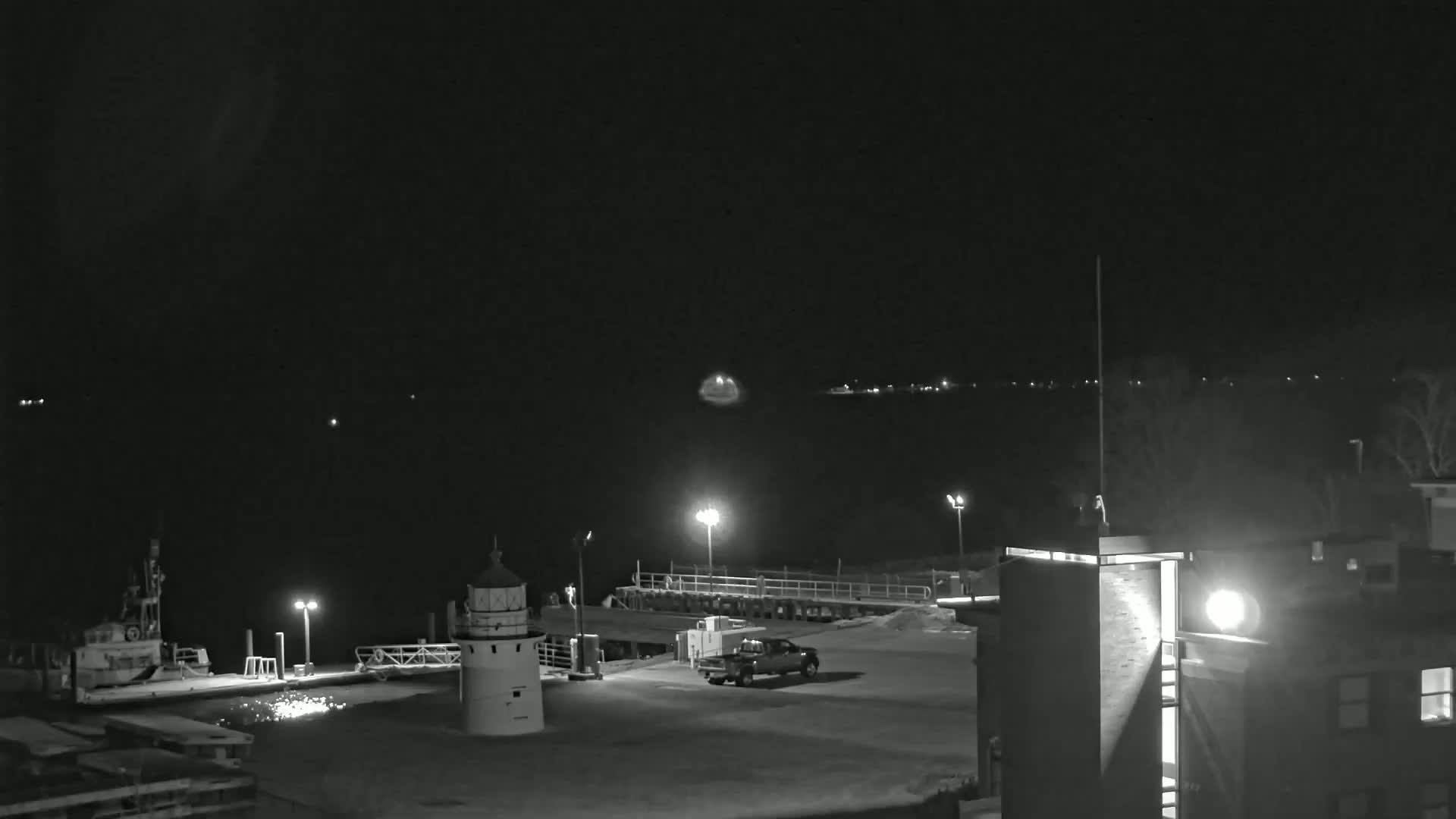 A nighttime, black-and-white view of a harbor with a lighthouse, a boat, a truck, and a building, under a clear sky.
