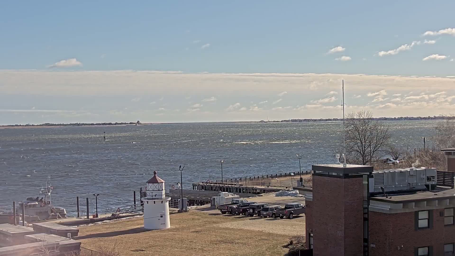 A small lighthouse stands on a grassy area near a dock and a body of water, under a mostly sunny sky with some clouds and a slight wind rippling the water's surface.