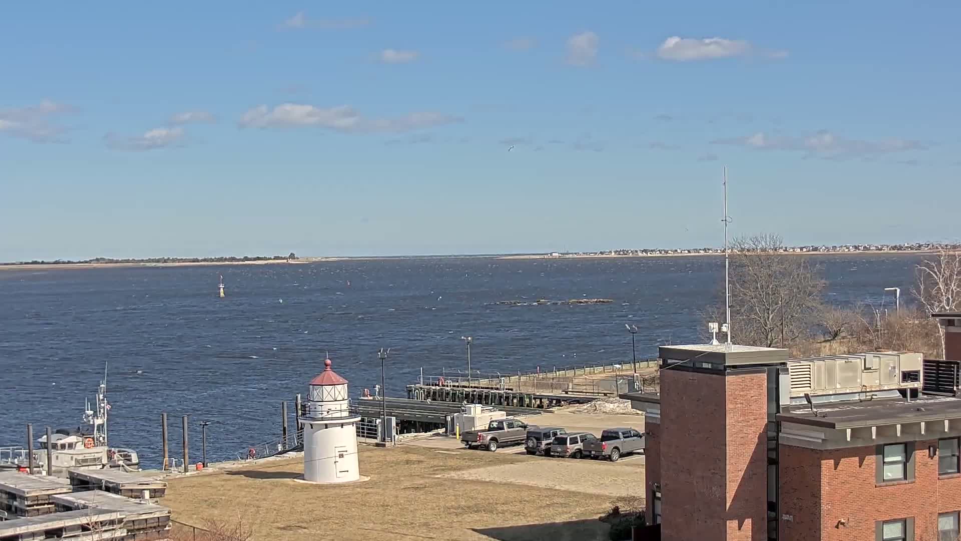 A small lighthouse stands on a grassy area next to a dock and a body of water under a mostly sunny sky with some scattered clouds, a larger body of land is visible across the water.