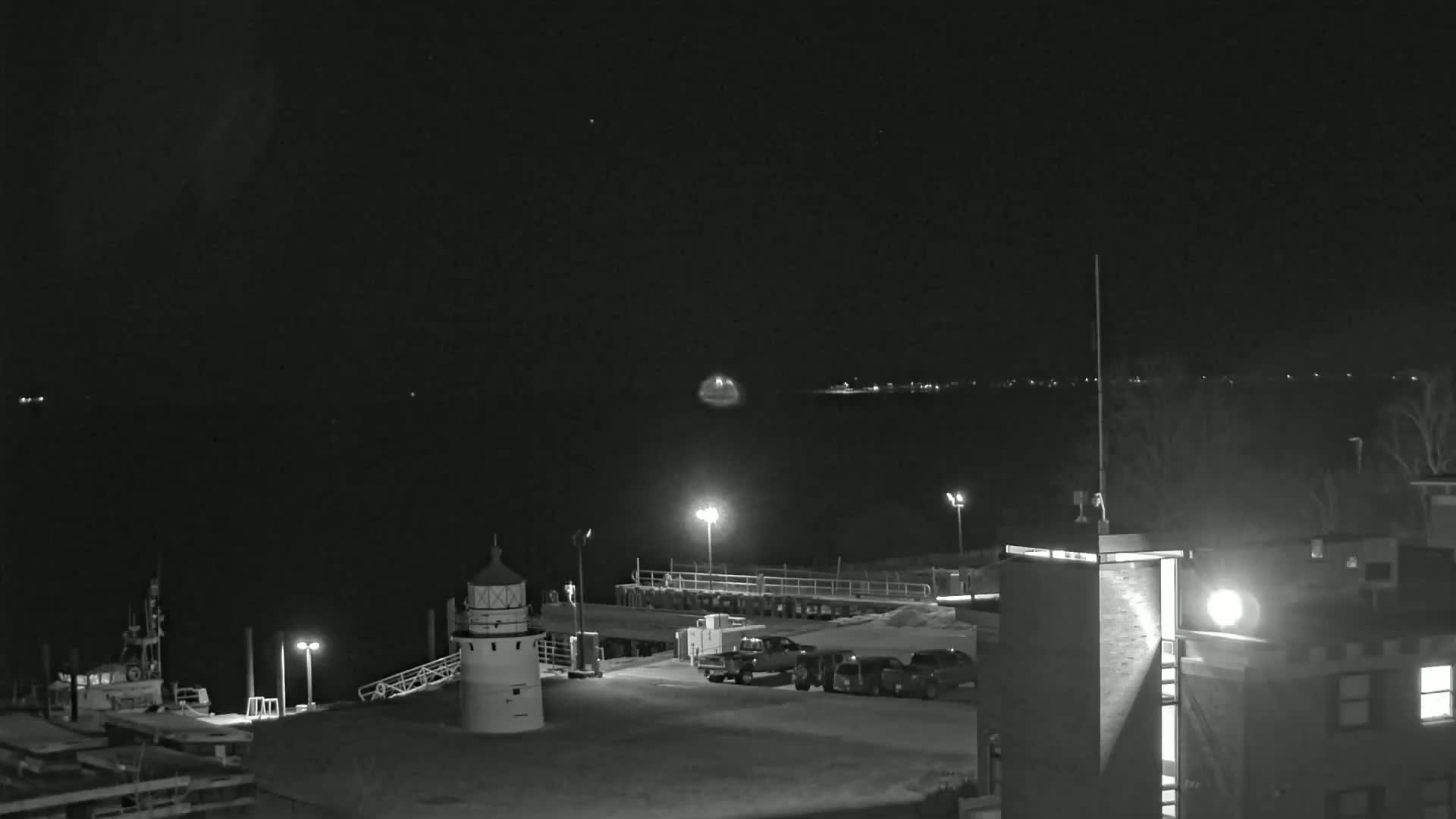 A nighttime, black-and-white view of a harbor with a lighthouse, several vehicles, and a building under a clear, dark sky.