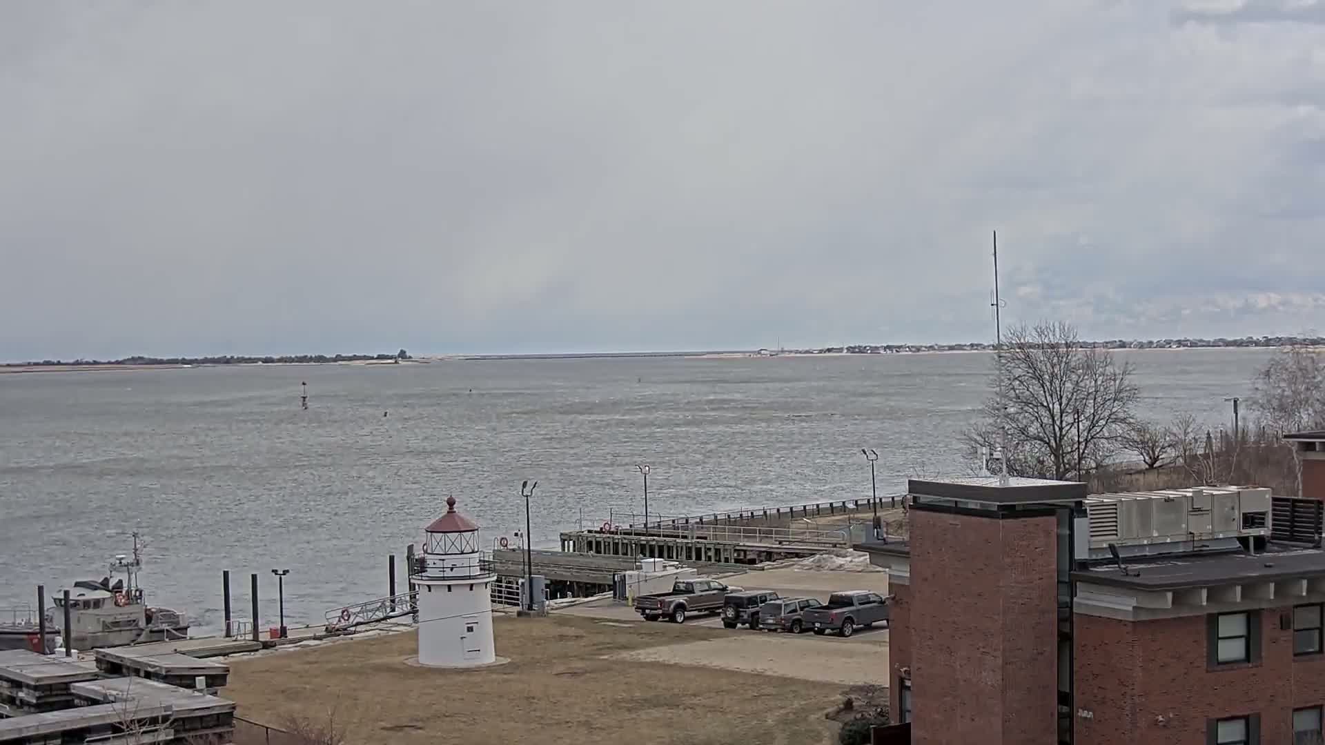 A small lighthouse stands near a dock and several vehicles, overlooking a body of water under an overcast sky.