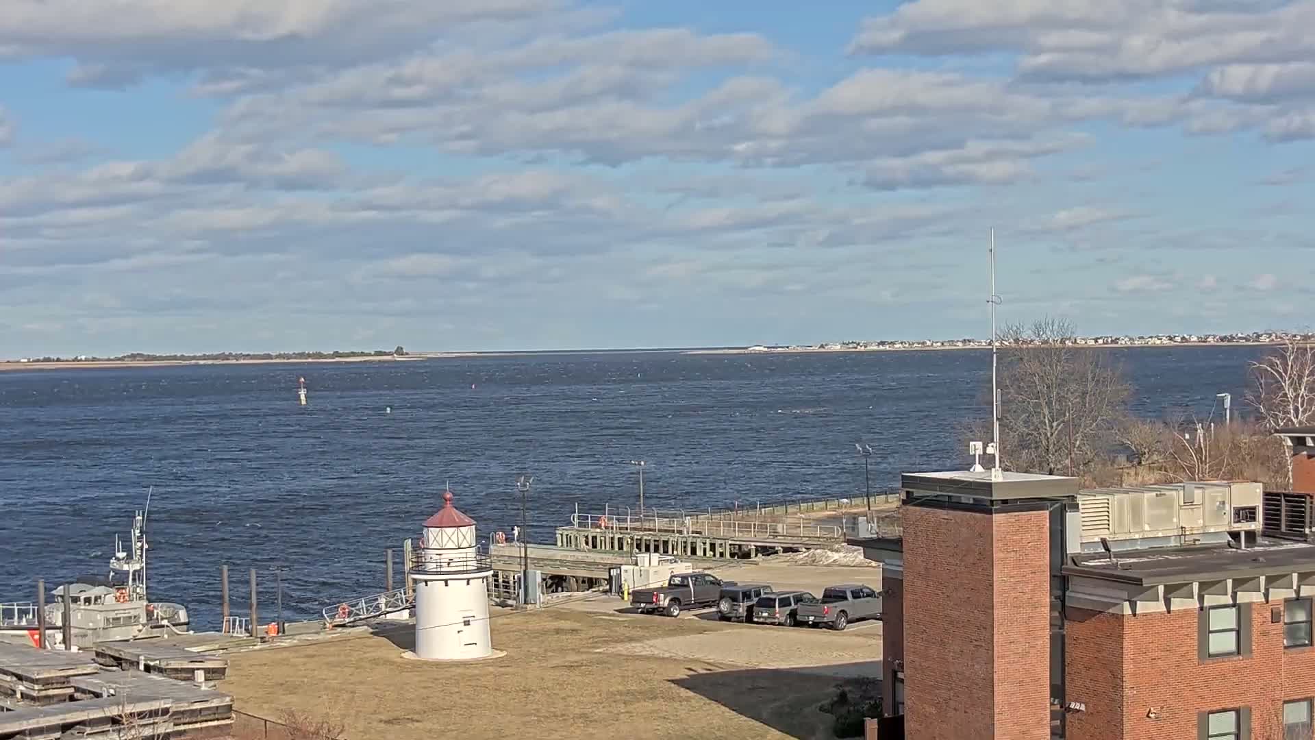 A white lighthouse stands on a grassy area near a dock with several vehicles and a small boat, overlooking a calm body of water under a partly cloudy sky.