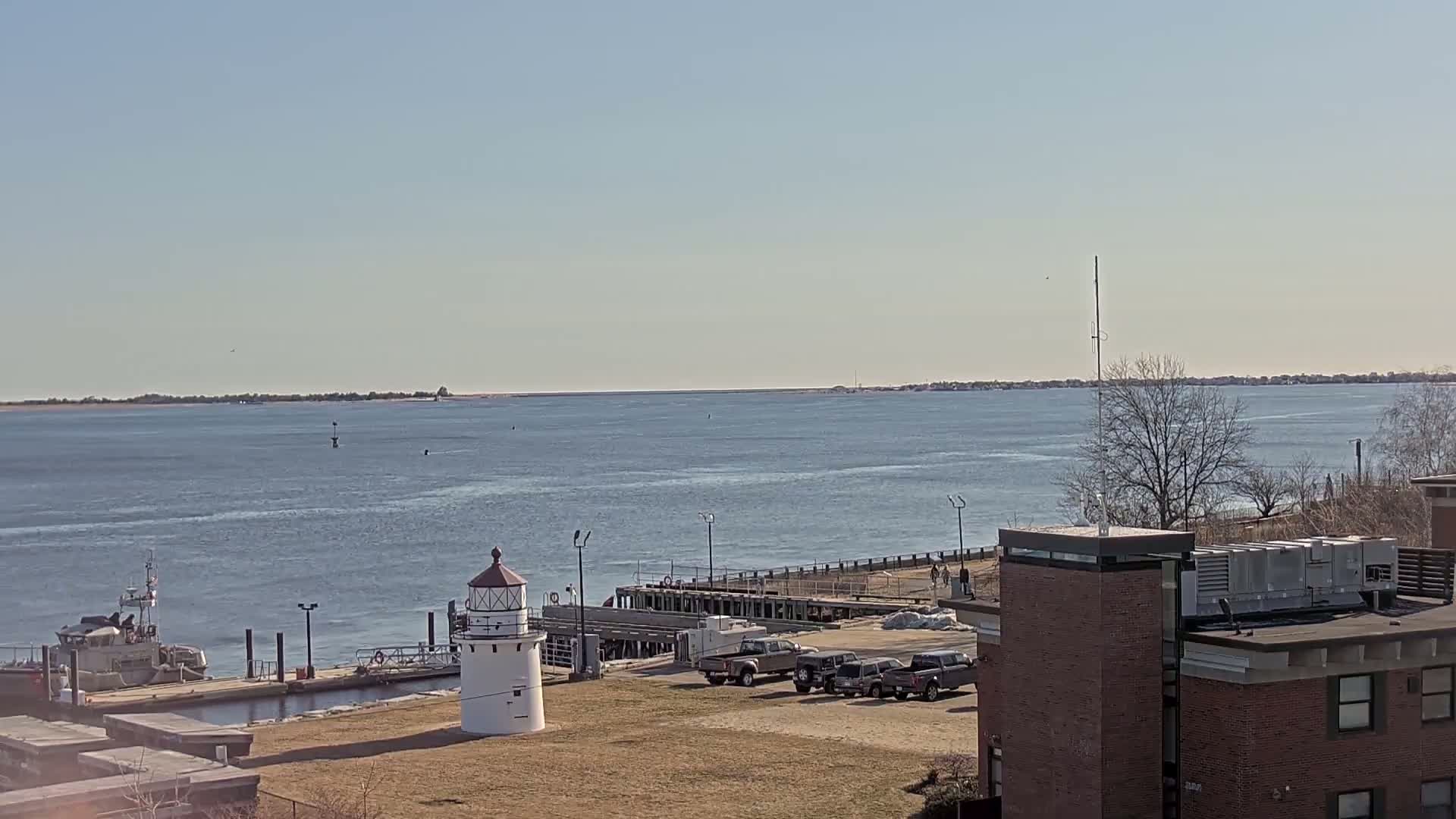 A small lighthouse stands near a harbor with a docked boat, several vehicles, and a brick building, all overlooking a calm body of water under a clear, sunny sky.