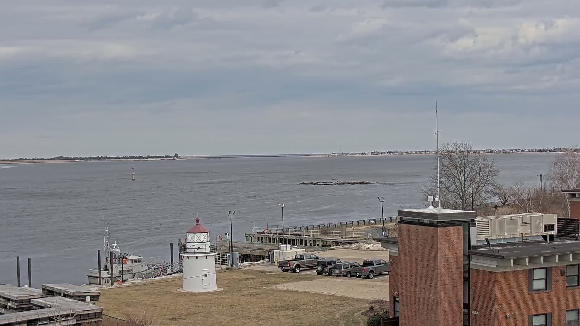 A small white lighthouse stands near a docked boat and several parked vehicles, overlooking a calm, gray body of water under a cloudy sky.