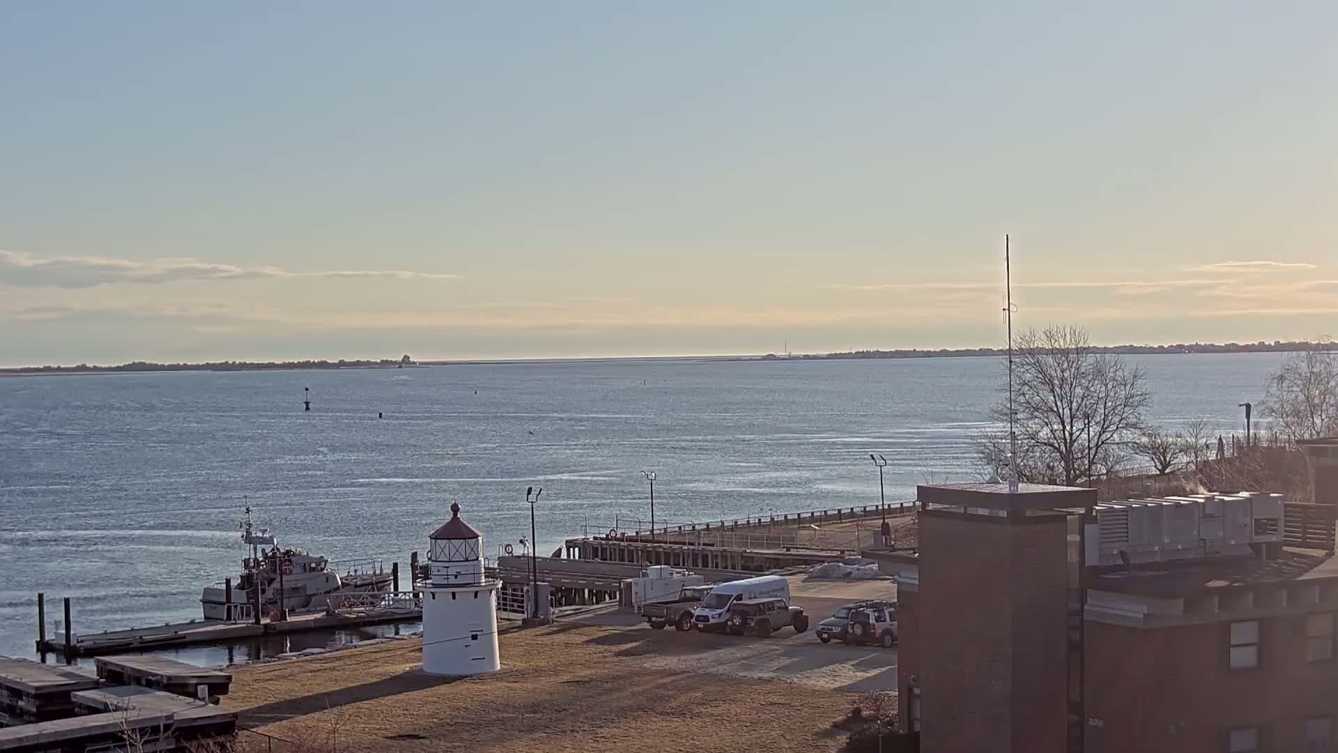 A calm, clear day reveals a harbor scene with a small lighthouse, docked boat, and several vehicles parked near a brick building overlooking a wide body of water.