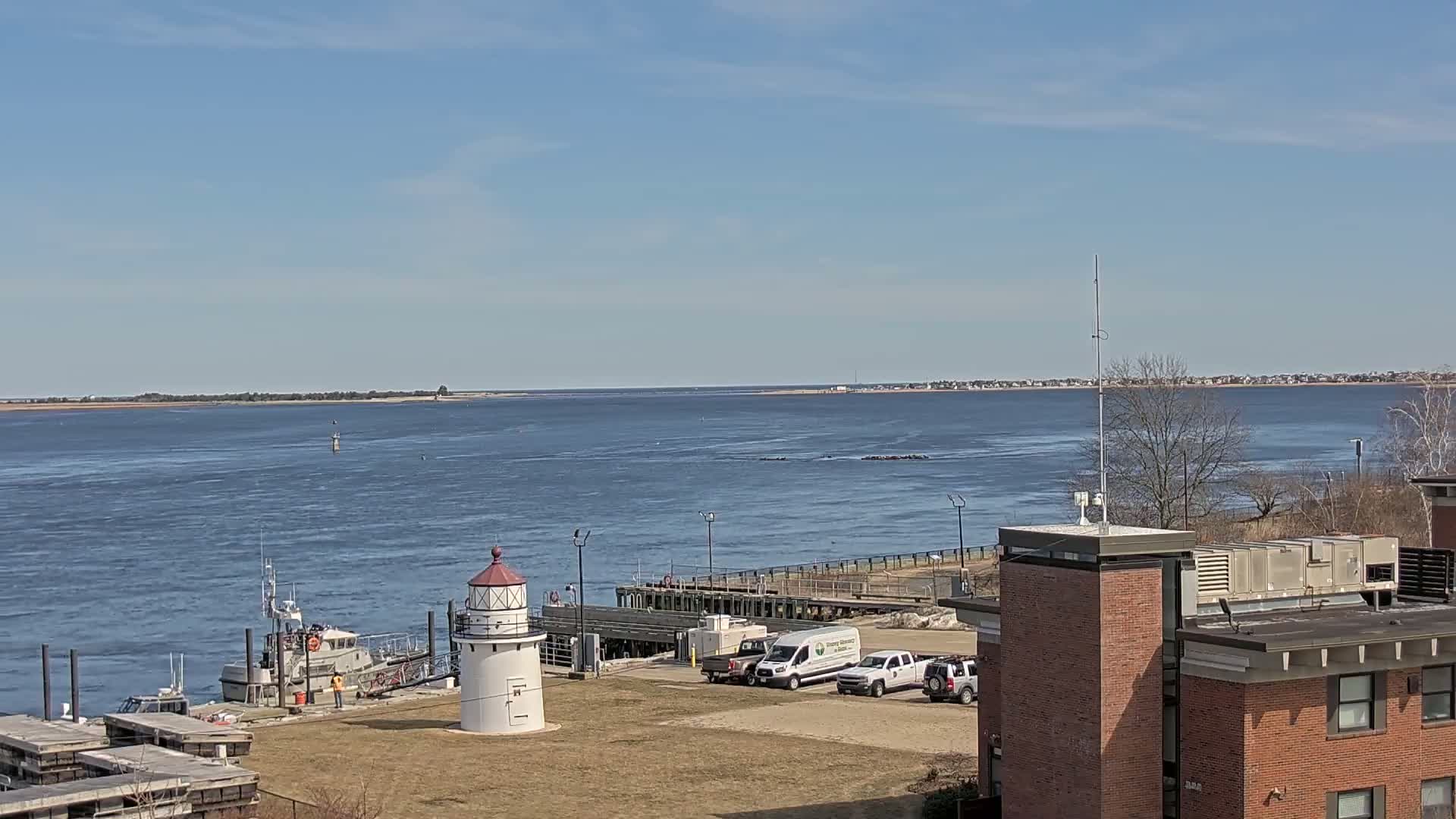 A small lighthouse stands near a dock with a boat and several vehicles, overlooking a calm body of water under a clear blue sky.