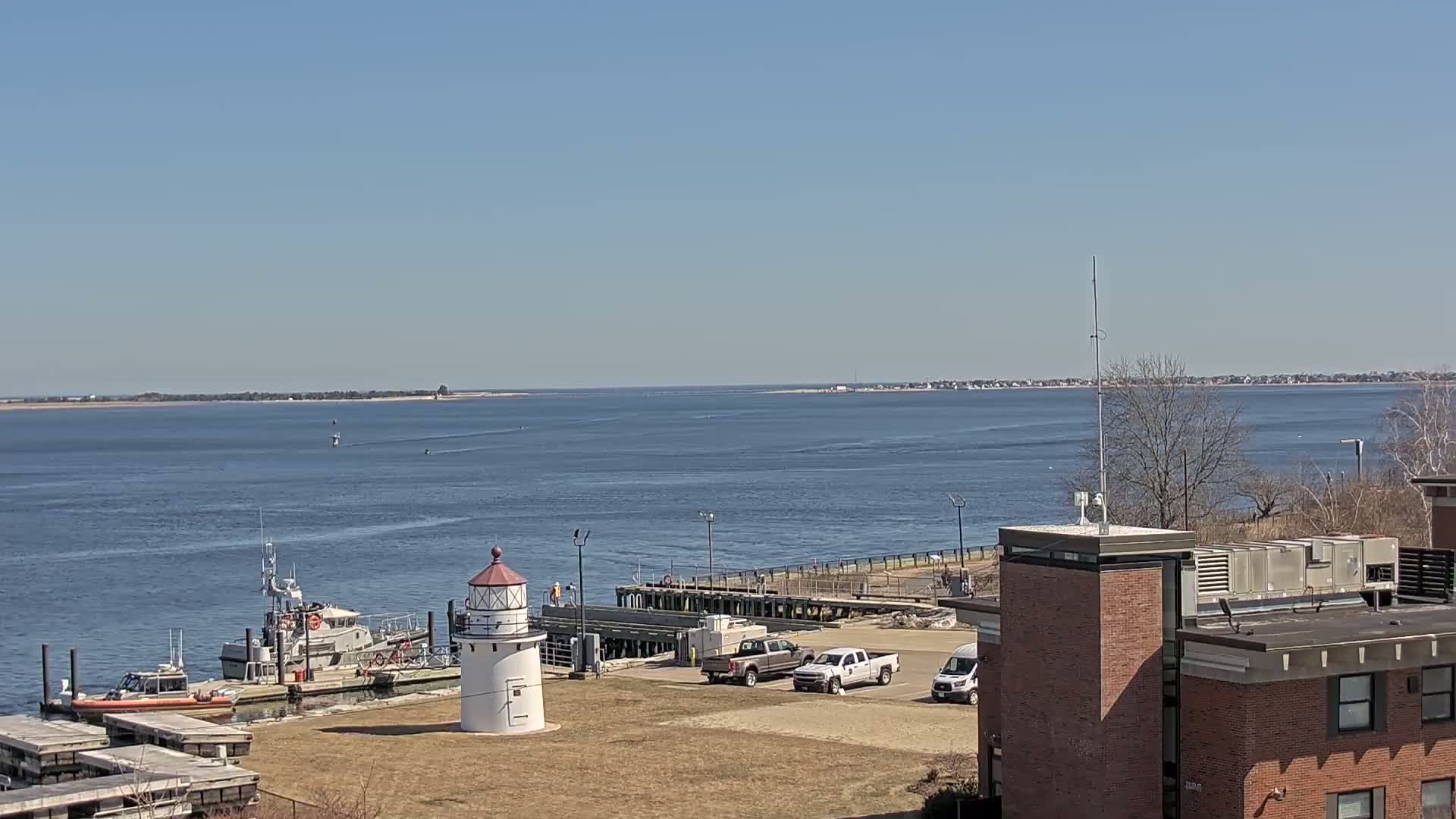 A small lighthouse stands near a dock with a Coast Guard boat and several other vehicles parked nearby, all overlooking a calm body of water under a clear blue sky.