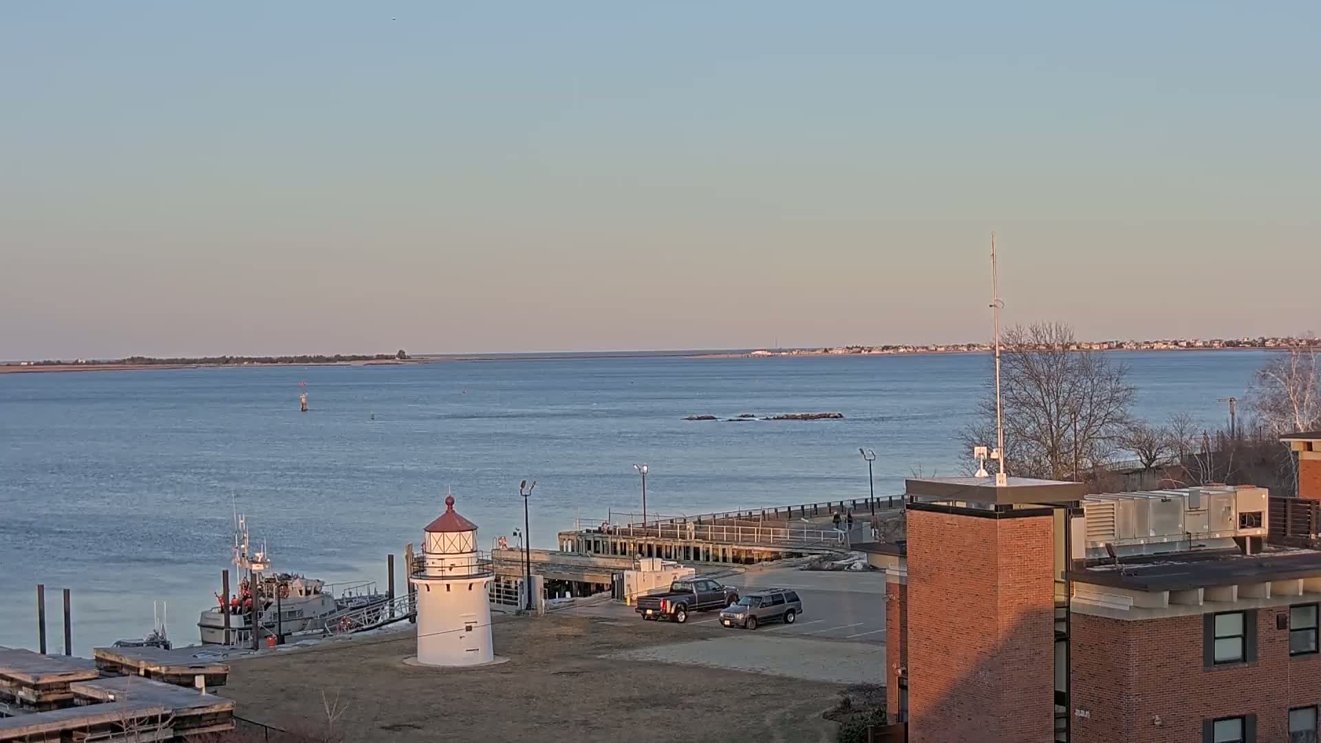 A small lighthouse stands near a docked boat on a calm body of water under a clear, pale sky, with a distant shoreline visible in the background and a brick building on the right.